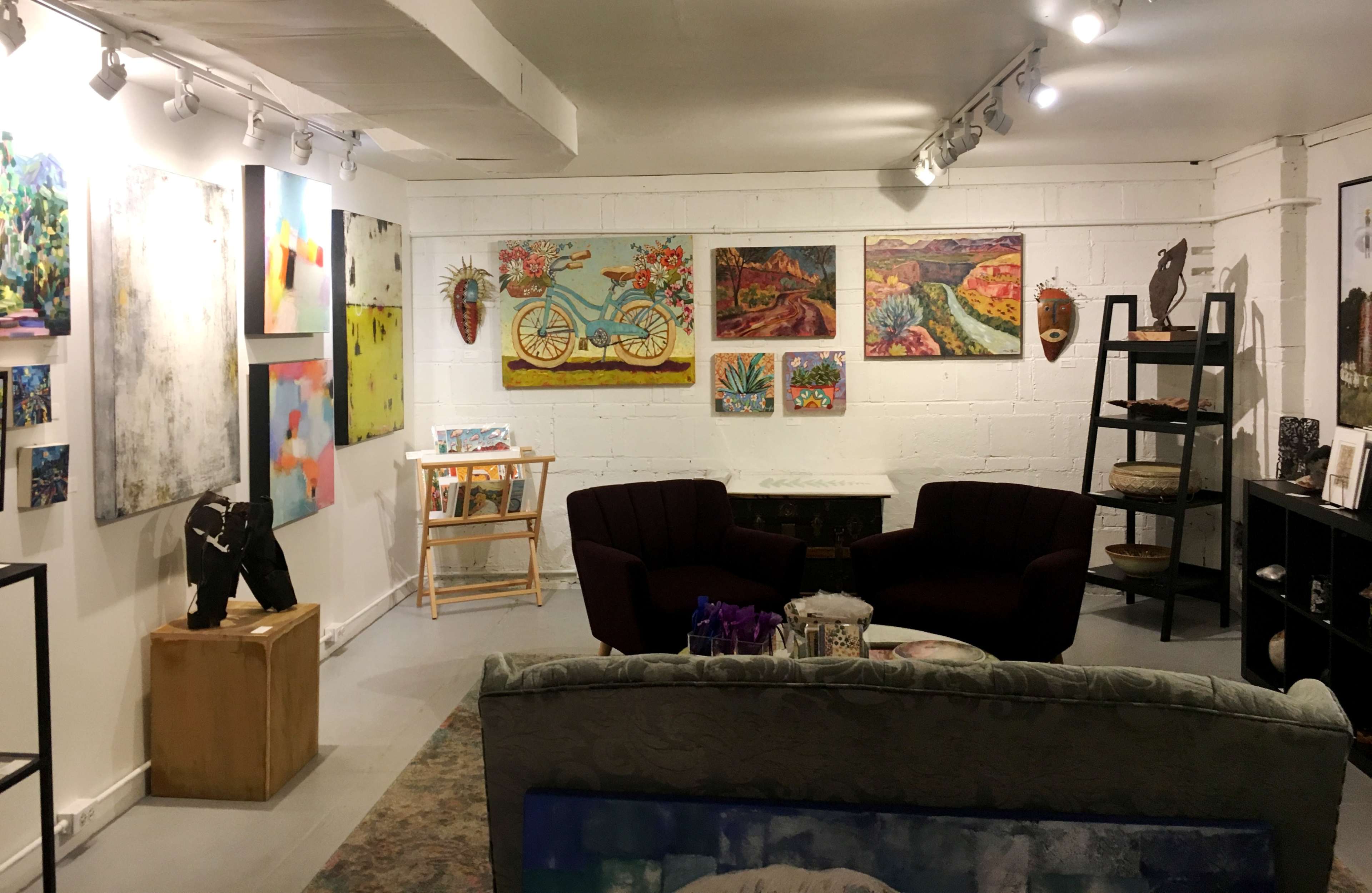 An art gallery featuring colorful paintings and sculptures displayed on the white walls and furniture of a well-lit room.
