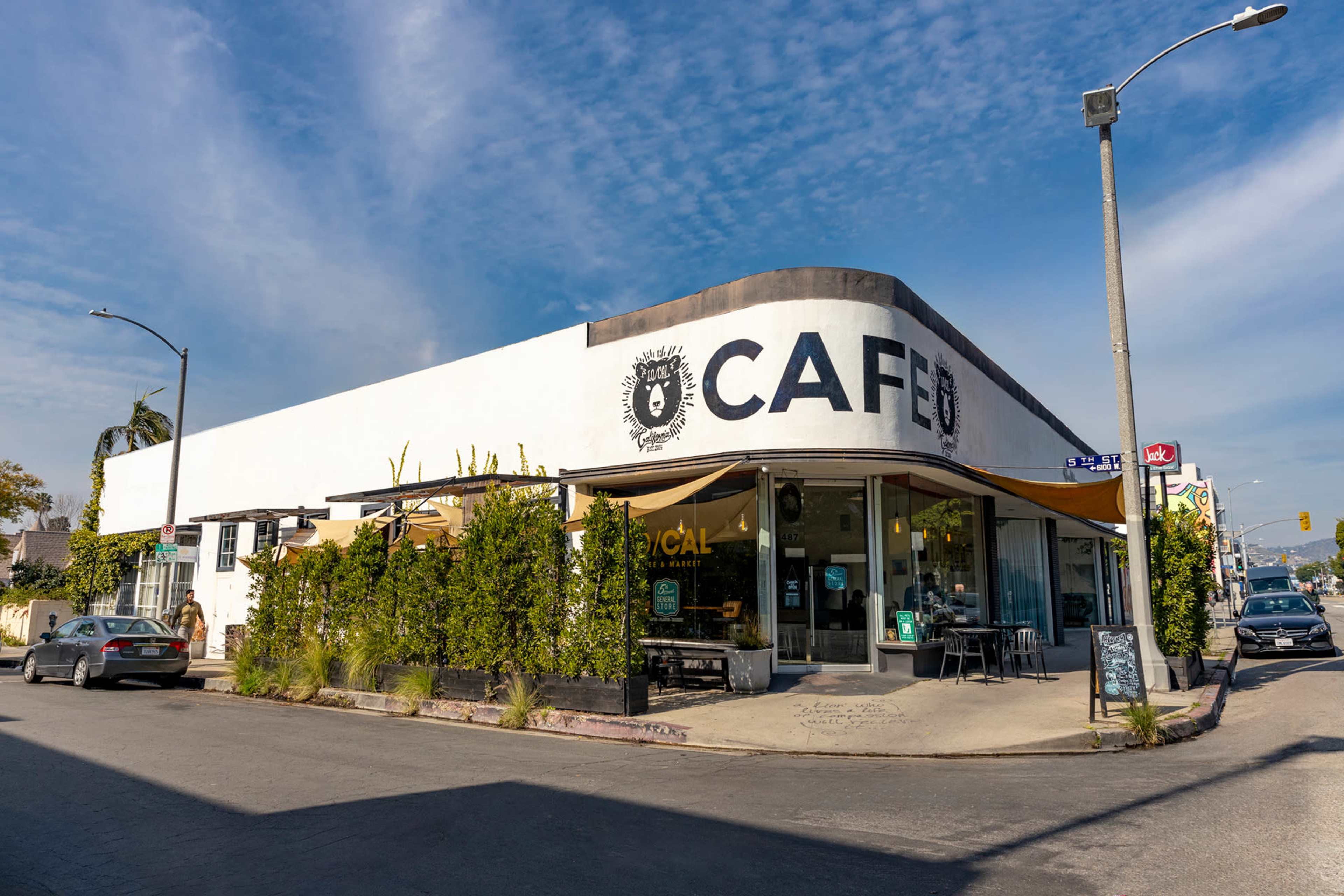 A corner cafe features a white exterior with large black lettering and outdoor seating surrounded by greenery.