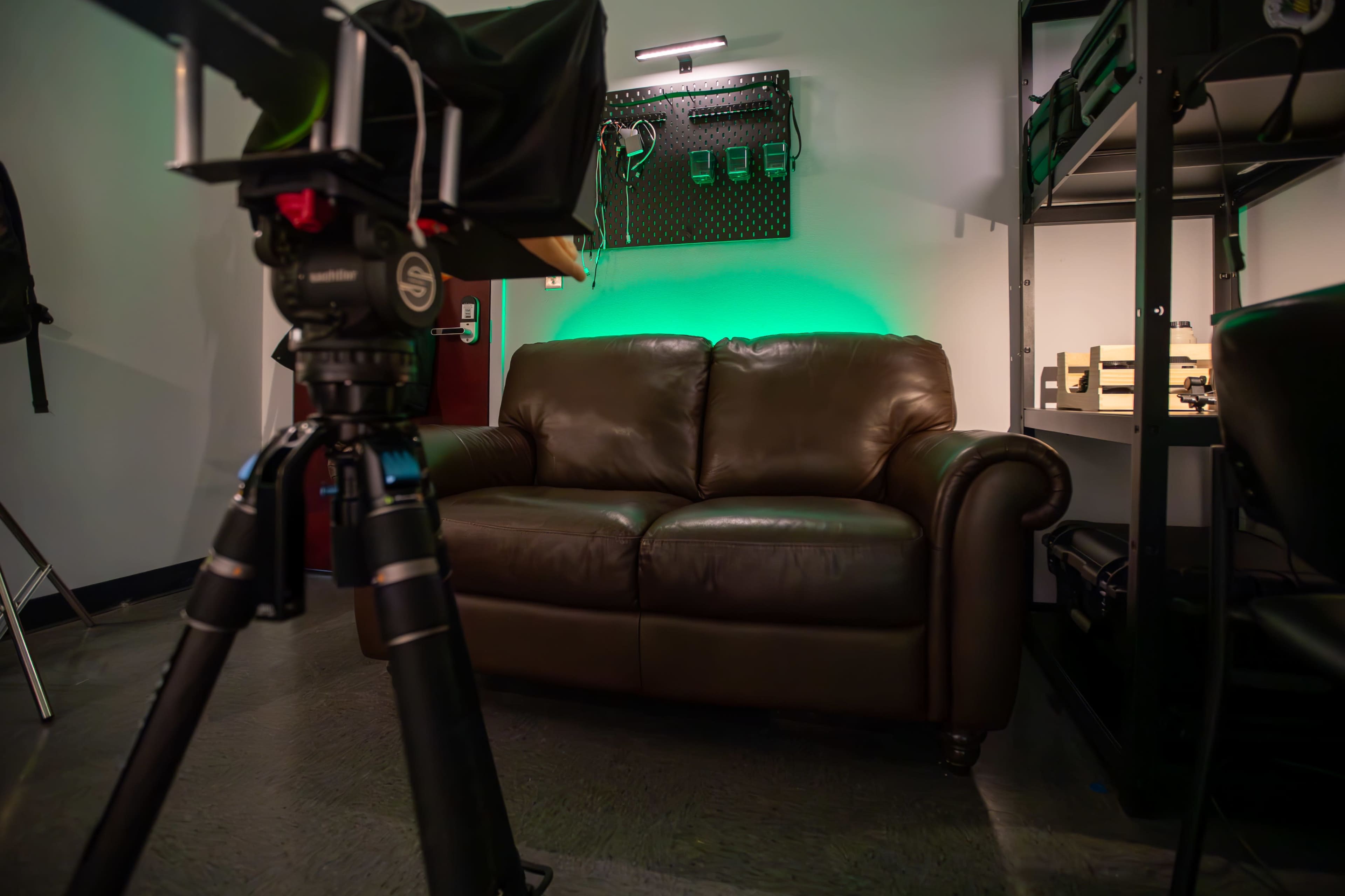The image shows a brown leather couch positioned in a room with a green-lit wall and various equipment on shelves in the background.
