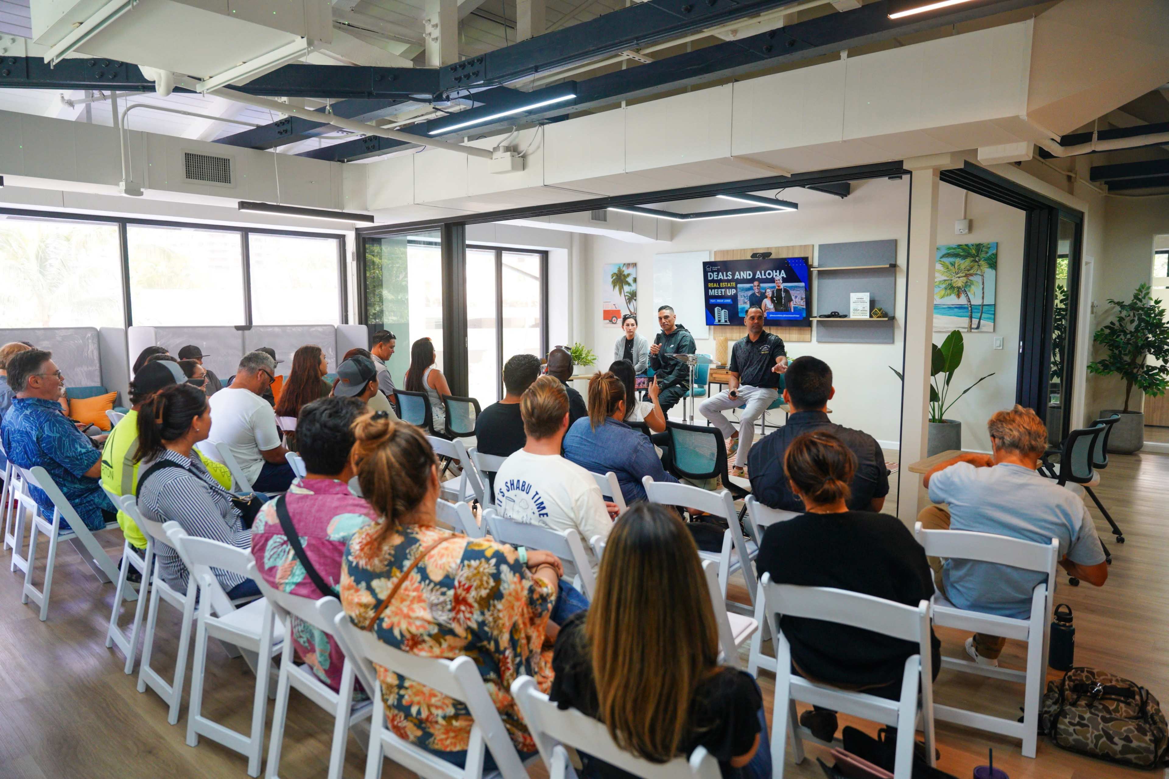 A group of people sits in a modern conference room, attentively listening to two speakers on a stage.