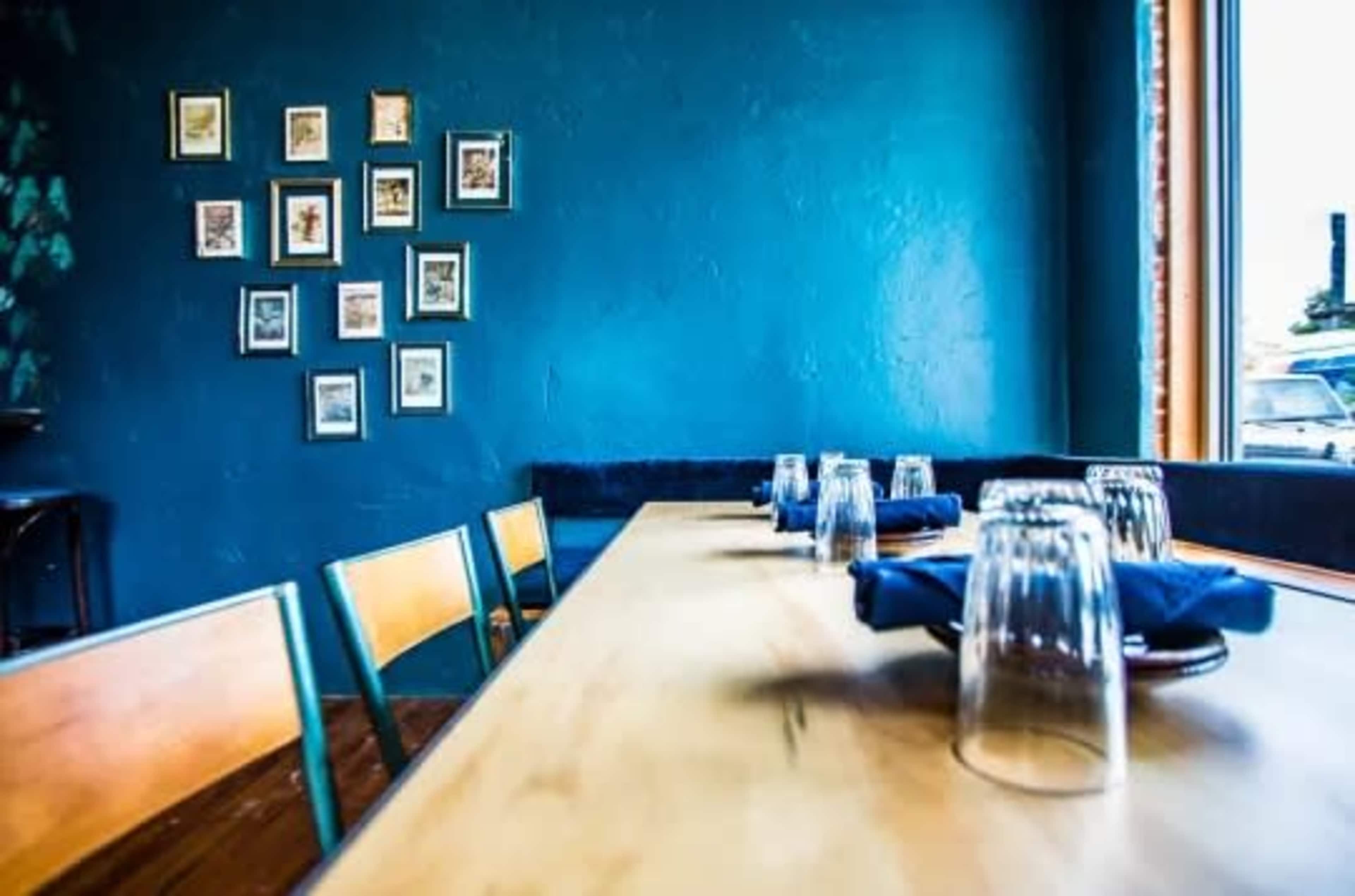 A long wooden table is set with glasses and blue napkins in a restaurant featuring a blue wall and framed pictures.