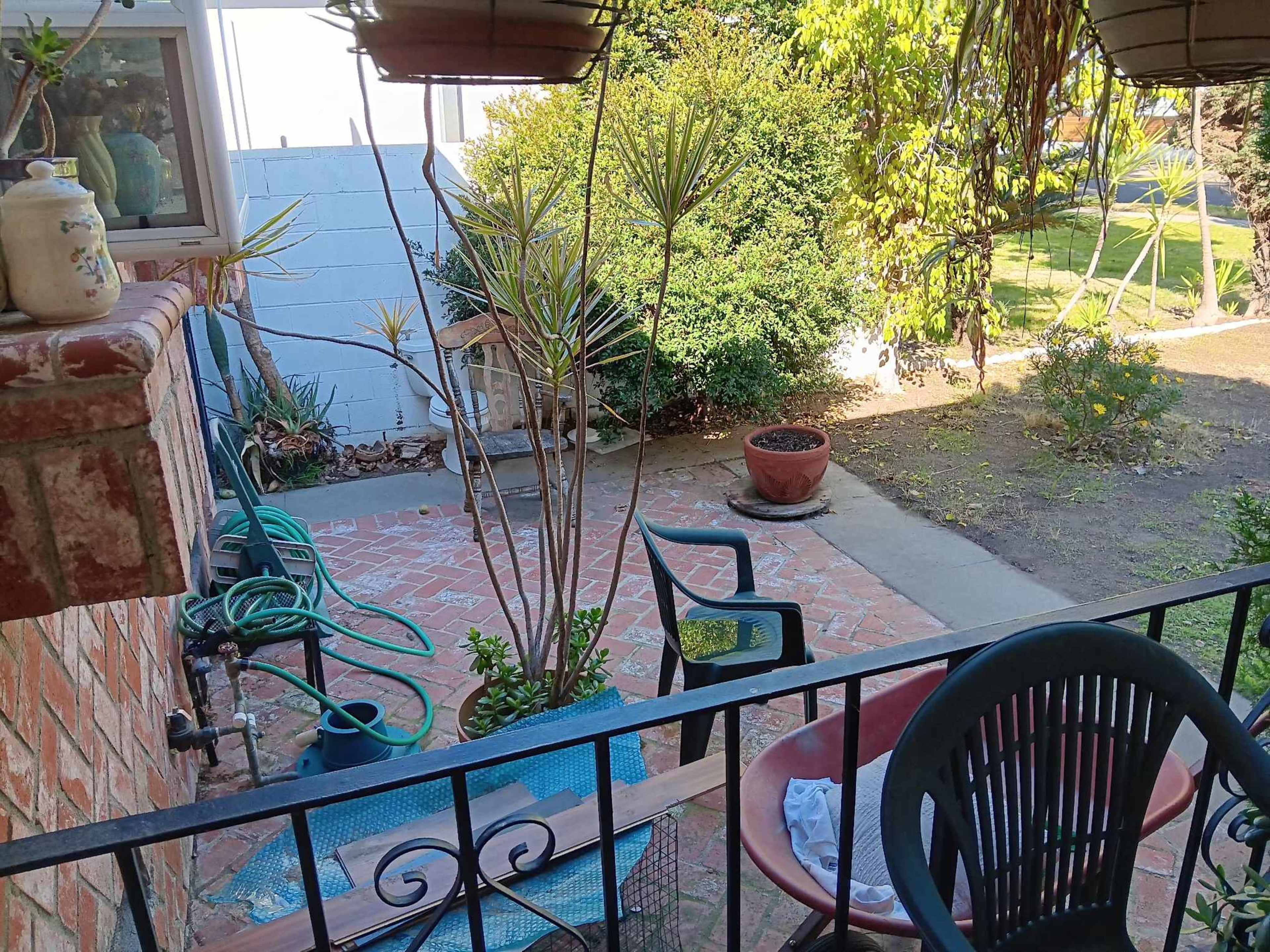A patio area is visible with potted plants, chairs, and a brick wall, framed by hanging planters and garden hoses.