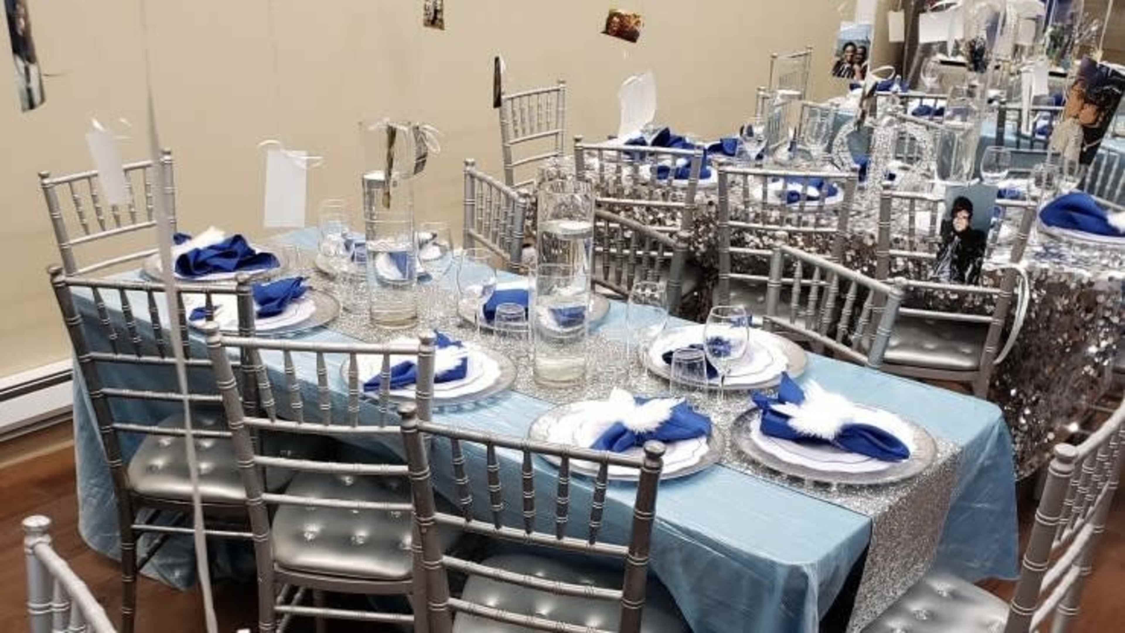 A banquet table is set with blue napkins, glassware, and silver decorations, arranged with chairs around it in a well-decorated room.