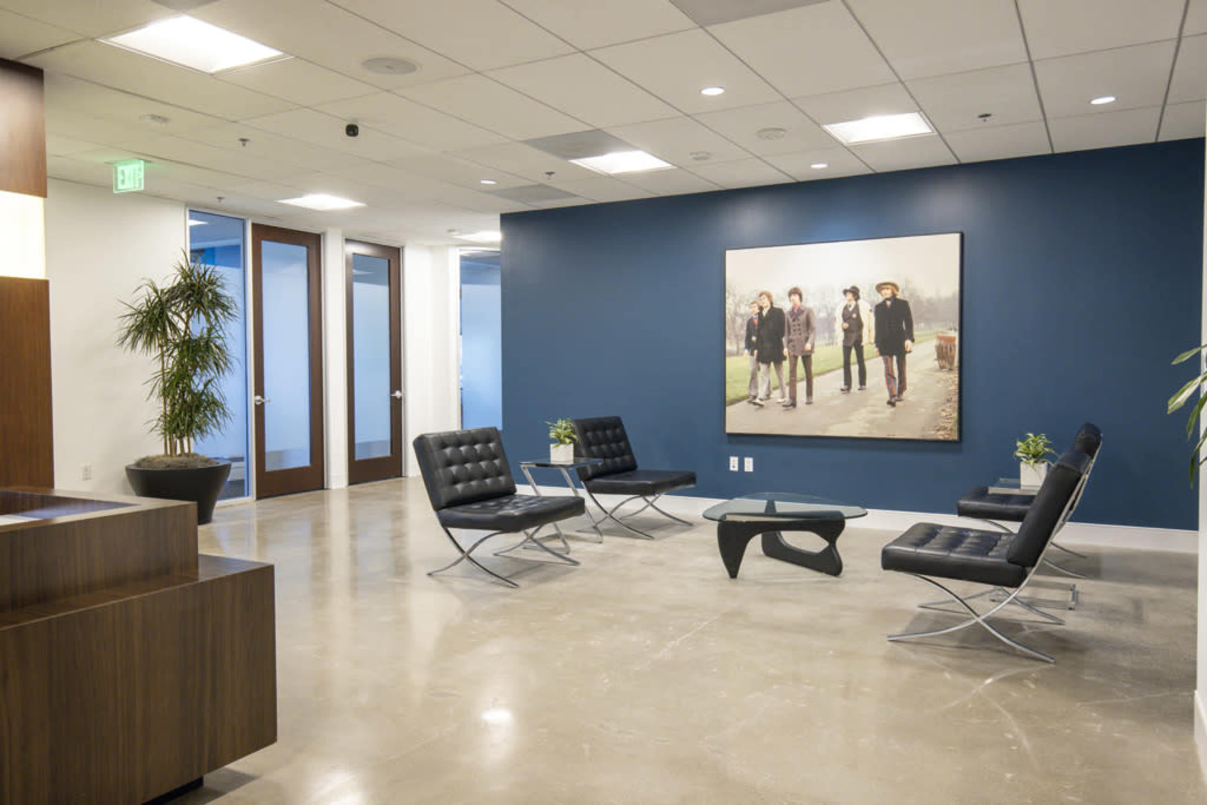 The image shows a modern office waiting area with four black chairs, a wooden reception desk, and a large artwork on the blue wall.