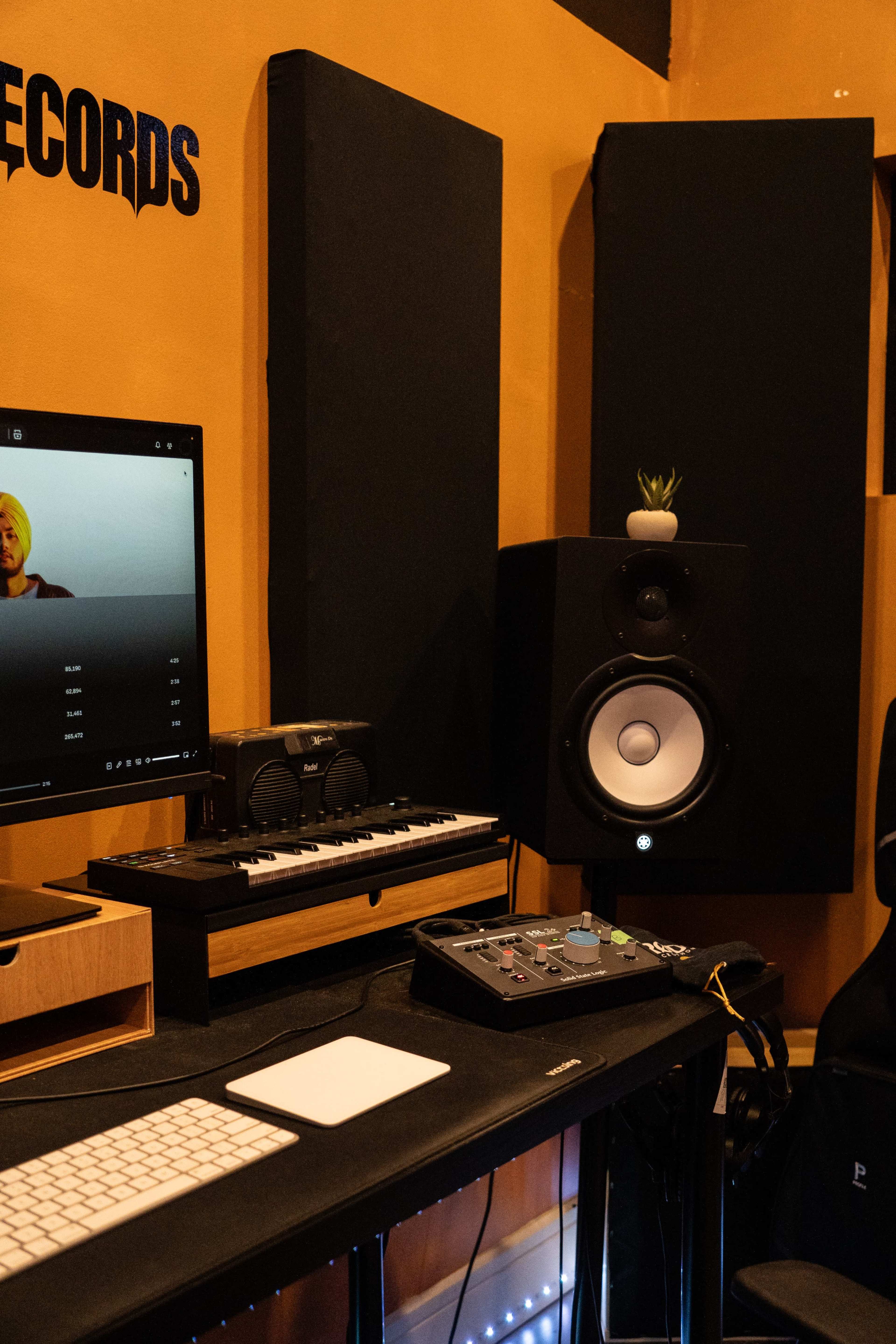 The image shows a music production studio setup with a computer, keyboard, audio interface, and speakers against an orange wall.