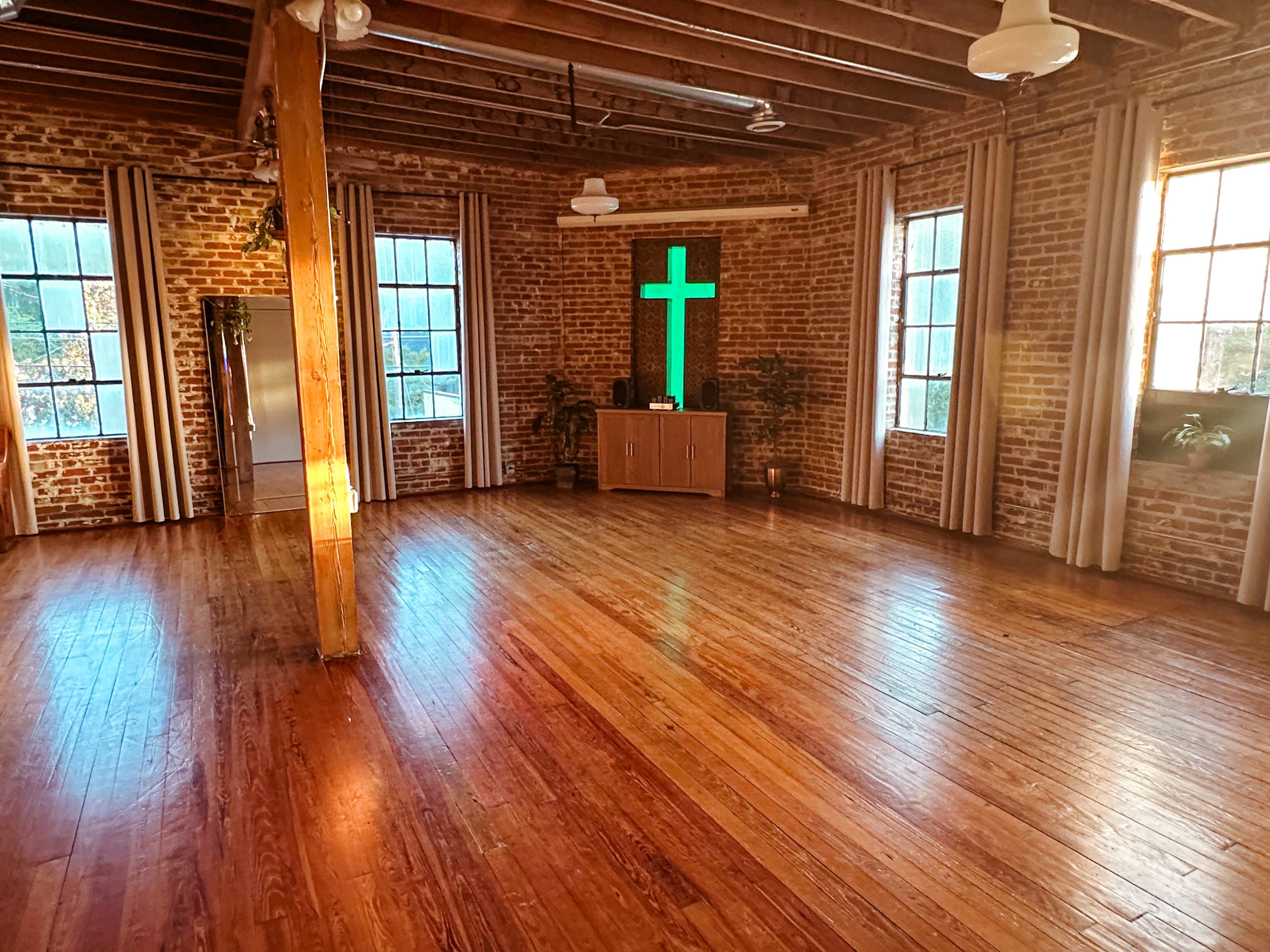 The image shows an empty room with wooden floors, exposed brick walls, large windows, and a green cross illuminated on one wall.
