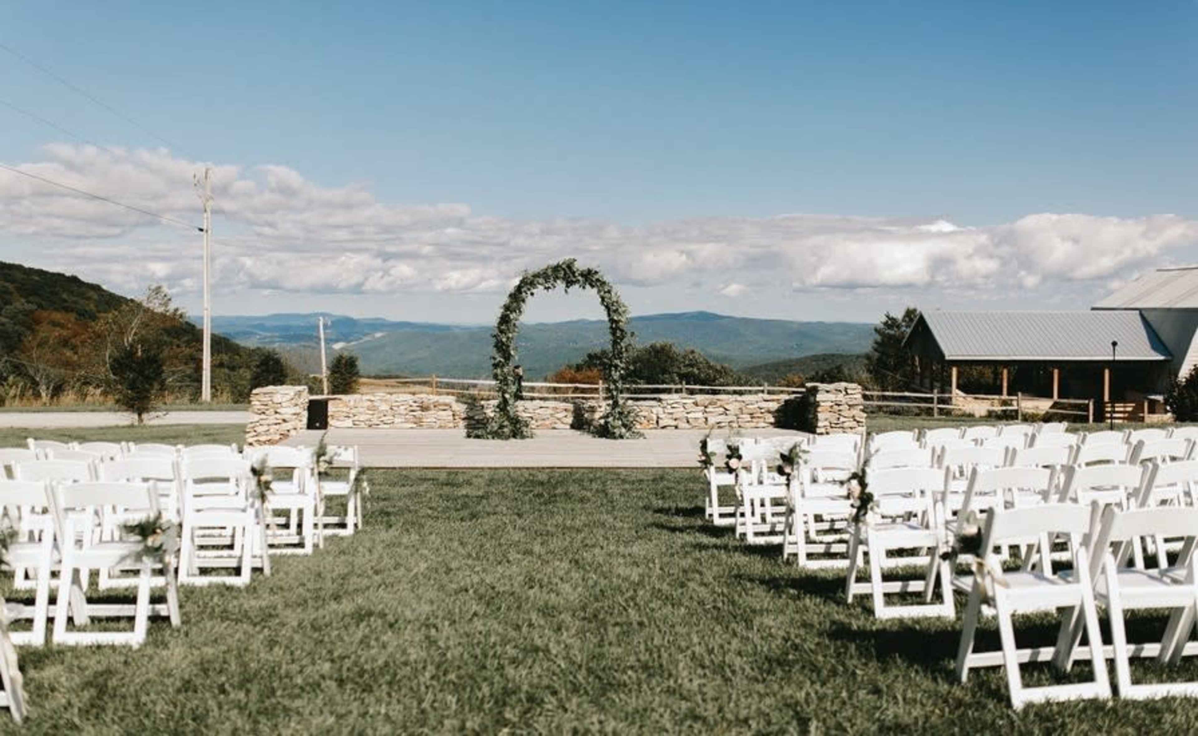 A scenic outdoor wedding venue features rows of white chairs facing a stone altar adorned with greenery, set against a backdrop of rolling hills and a blue sky.