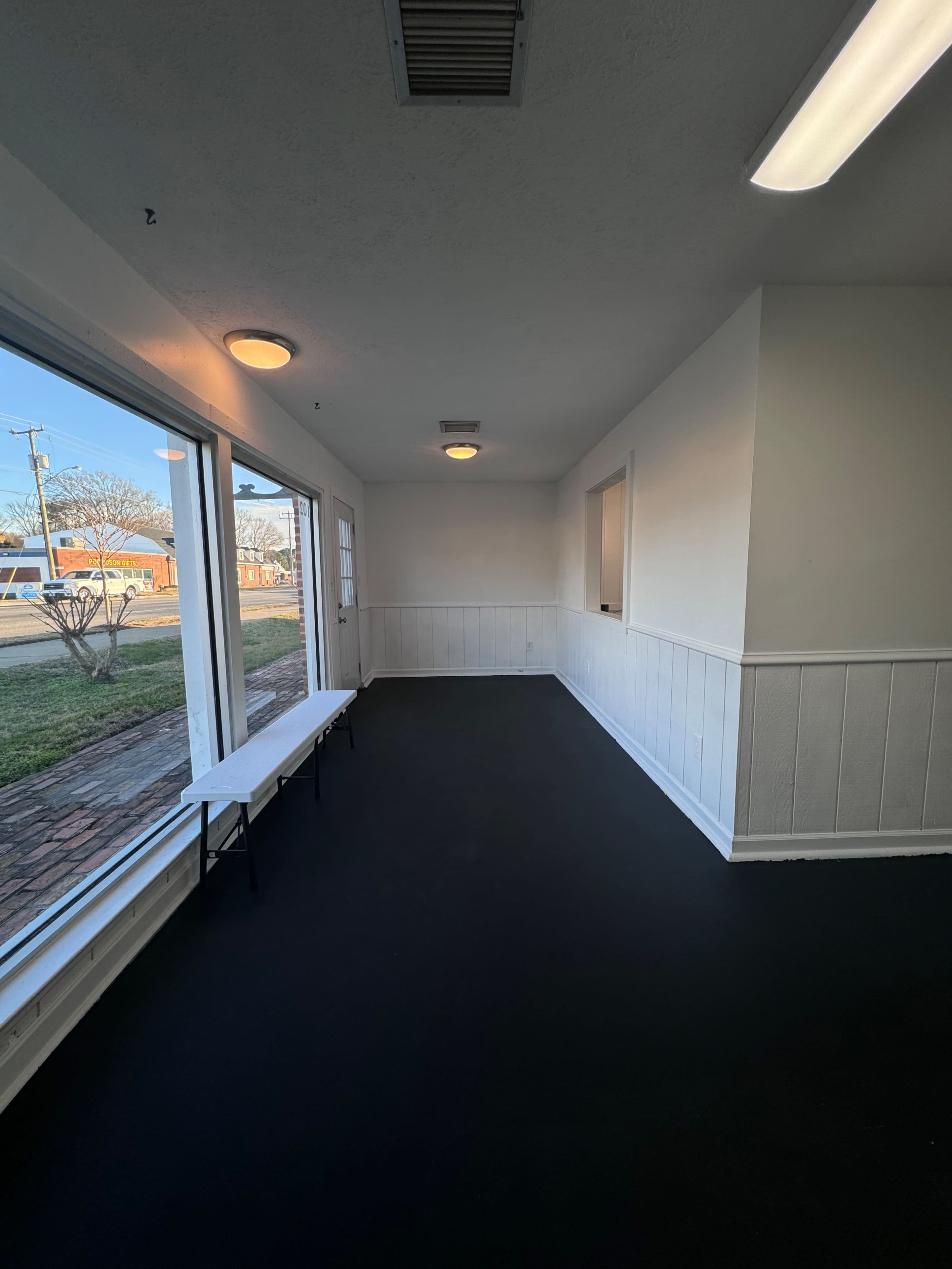 A sparsely furnished room with black flooring, white walls, and large windows allowing natural light to enter.