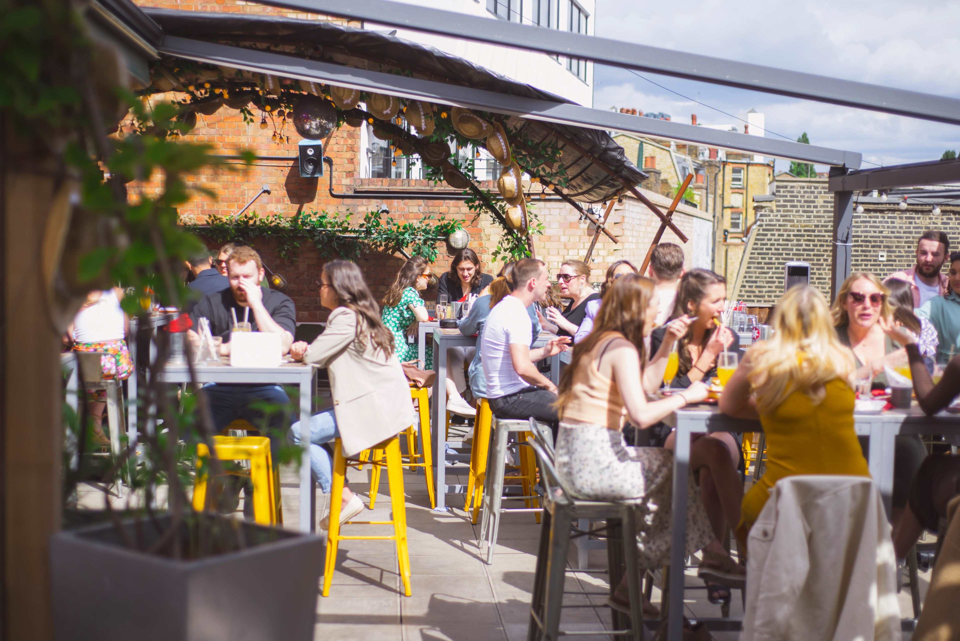 A bustling rooftop bar features groups of people seated at tables, enjoying drinks and conversation under a partially covered area.
