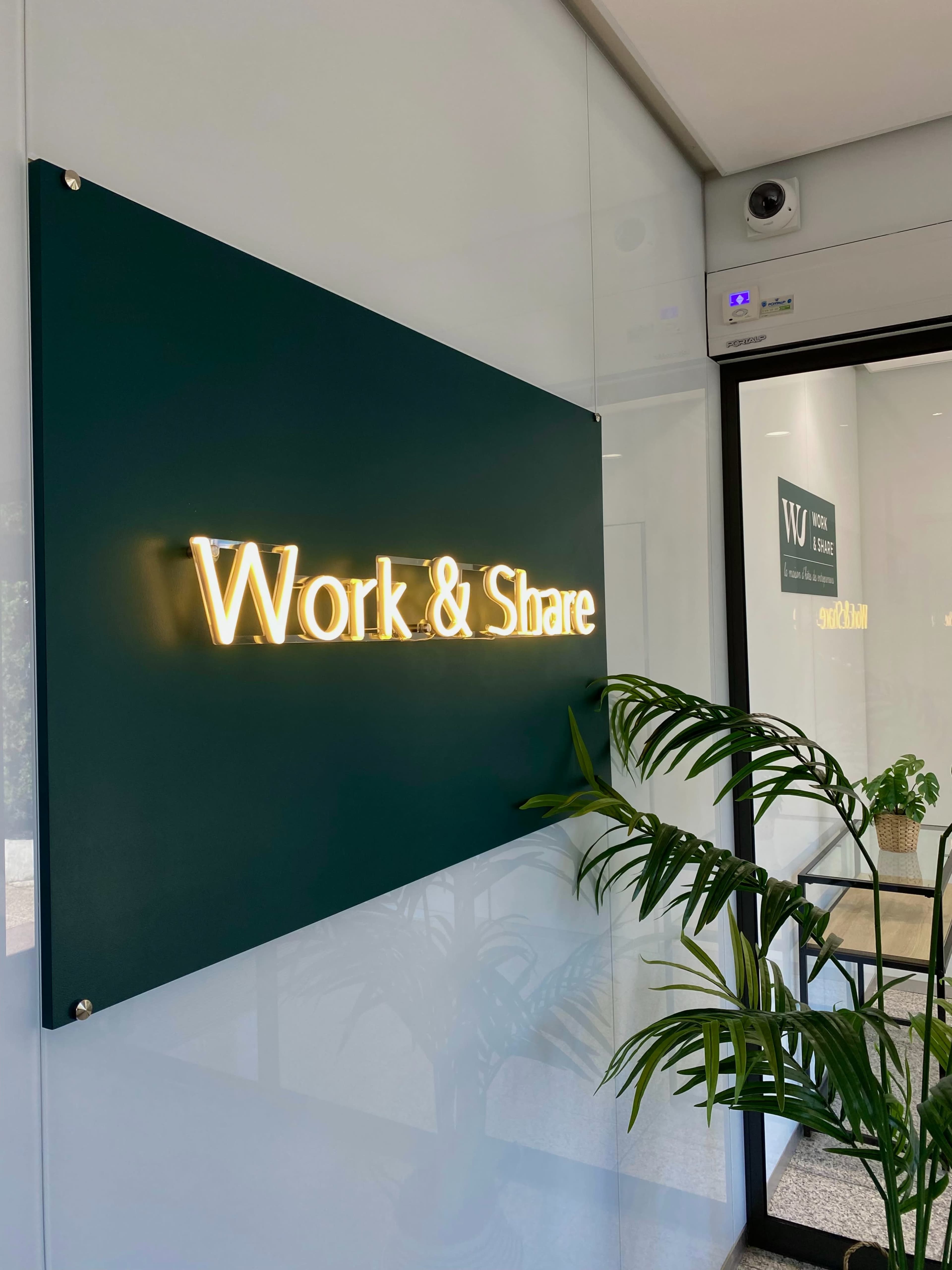 A illuminated sign that reads "Work & Share" mounted on a green background, with a potted plant in the foreground.