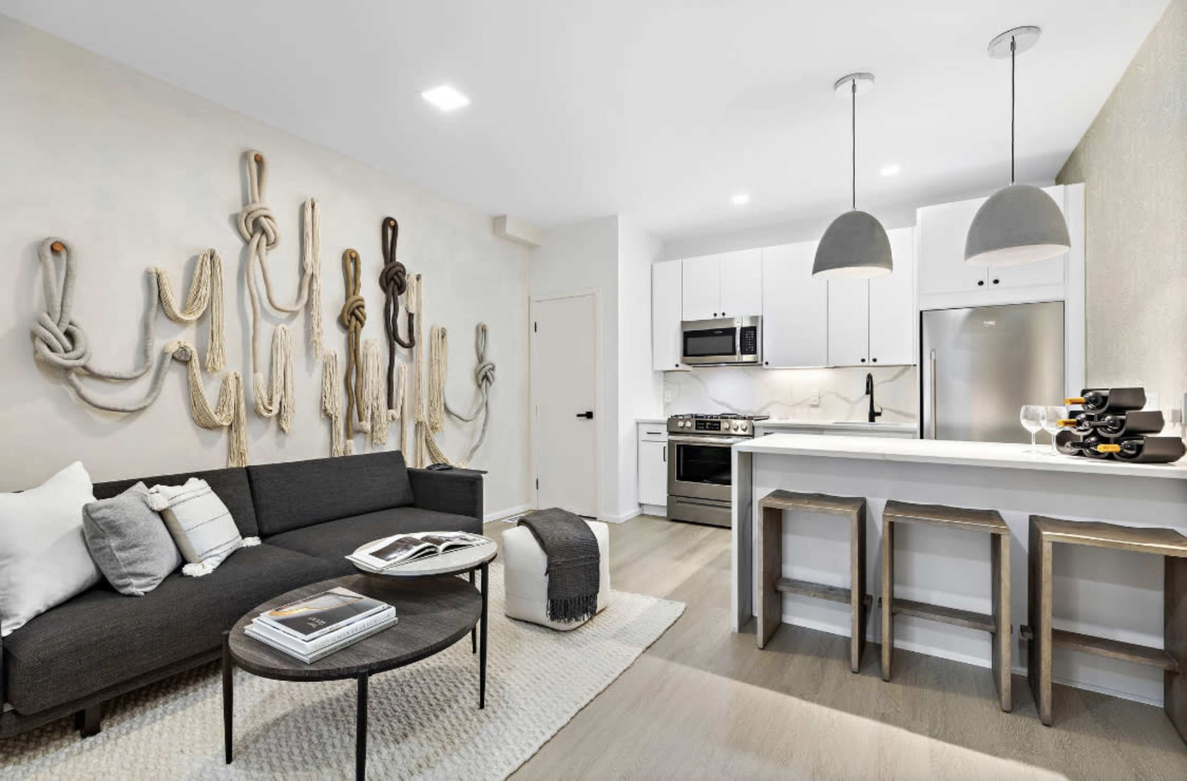 A modern apartment interior featuring a dark sofa, a round coffee table, and a kitchen area with white cabinetry and stainless steel appliances.