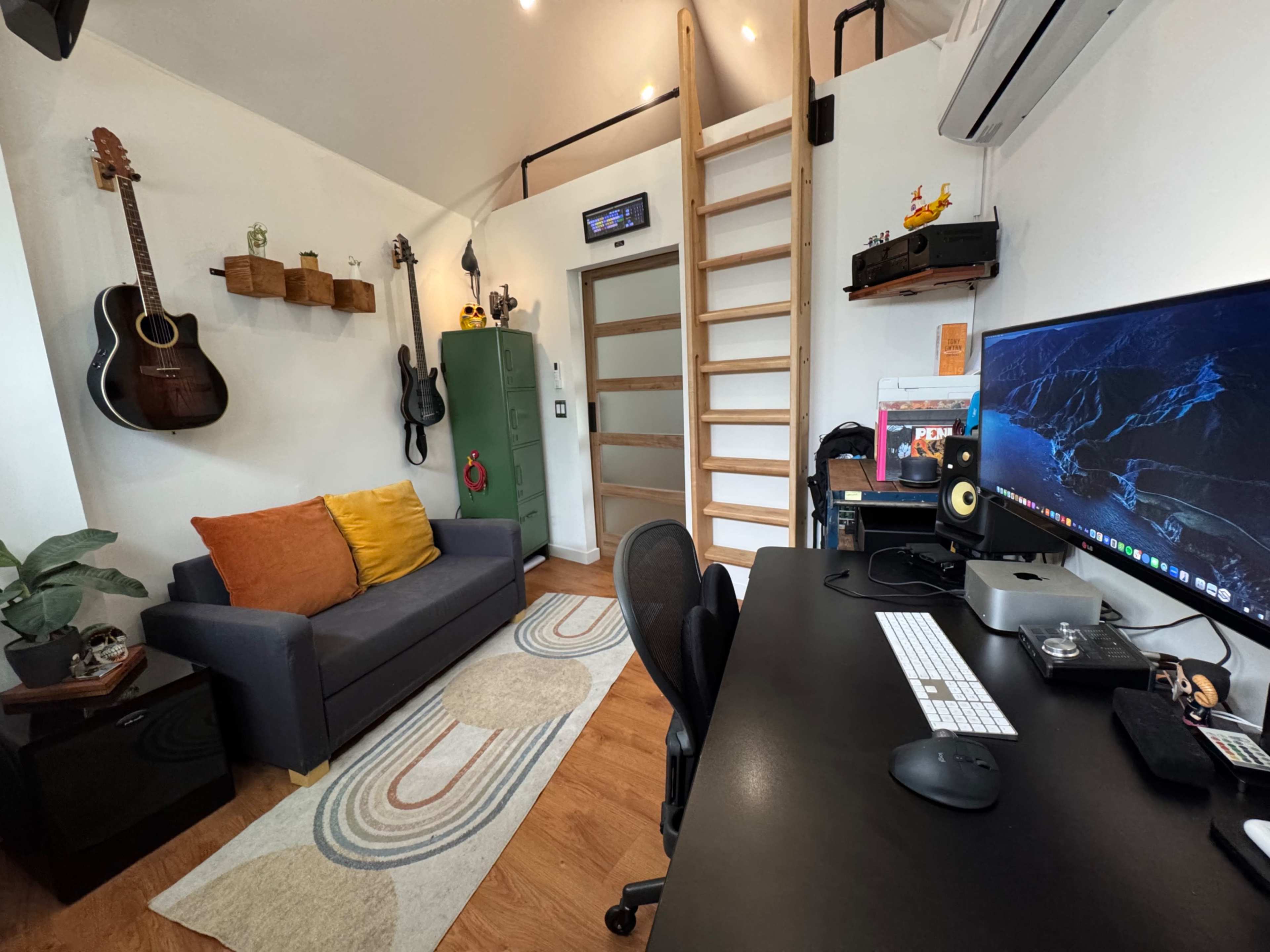 The image shows a small home office with a desk, computer setup, a gray sofa, and decorative items on the wall.