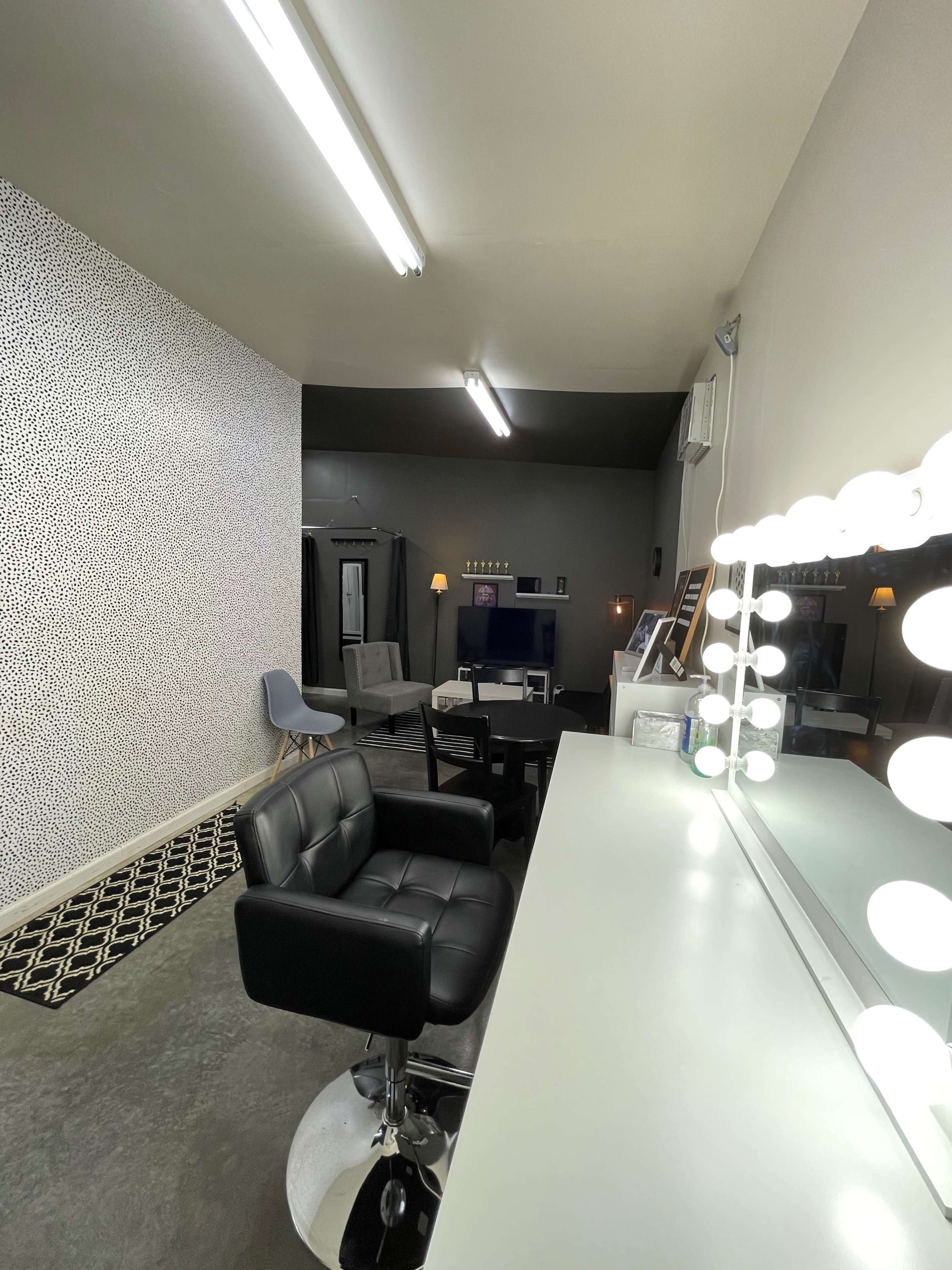 The image shows a modern room designed as a salon or makeup studio, featuring a black chair in front of a mirror illuminated by bright bulbs, with decorative walls and seating areas in the background.