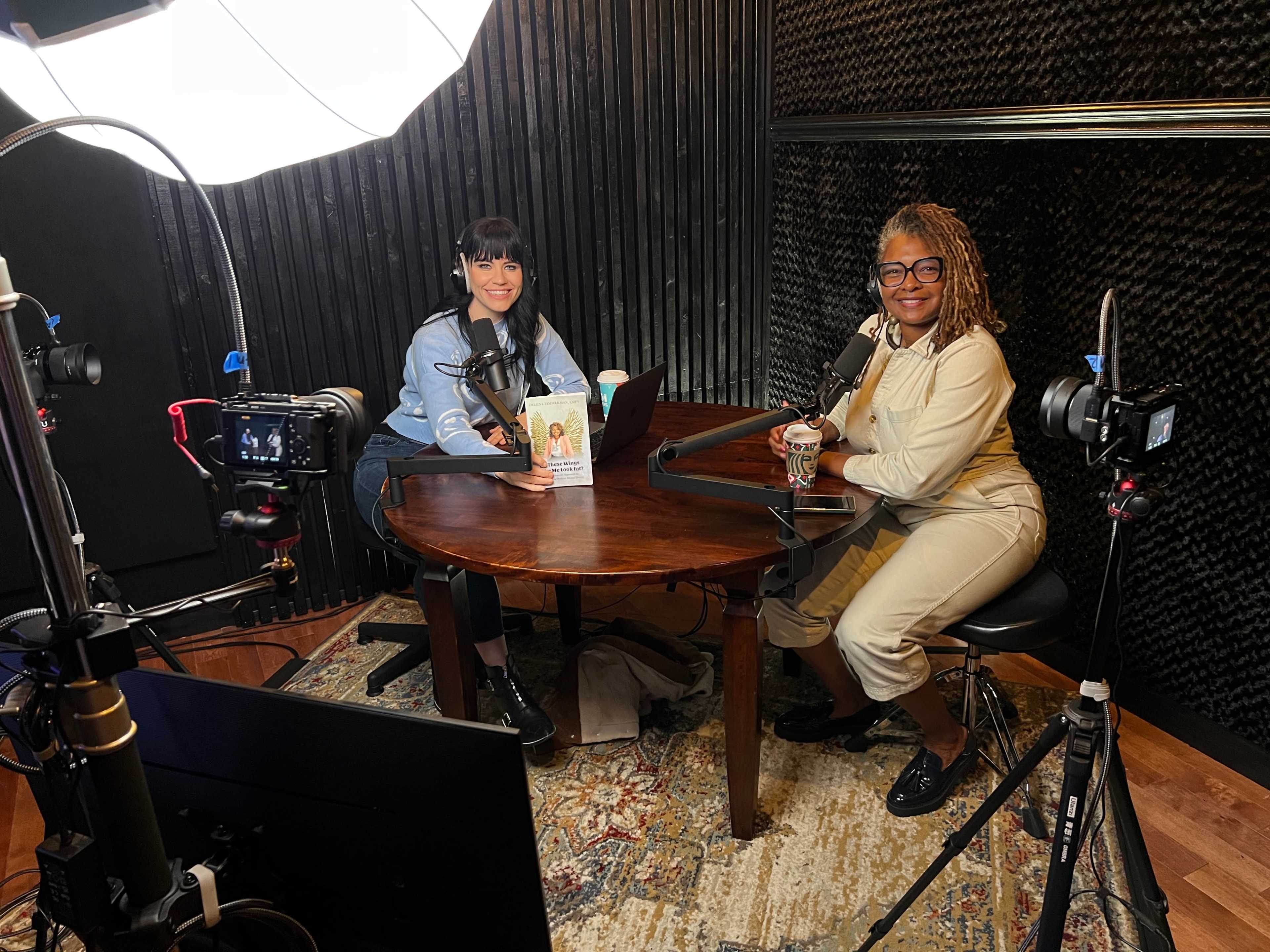 Two women are seated at a table in a podcast studio, equipped with microphones and camera equipment, with a large softbox light overhead.