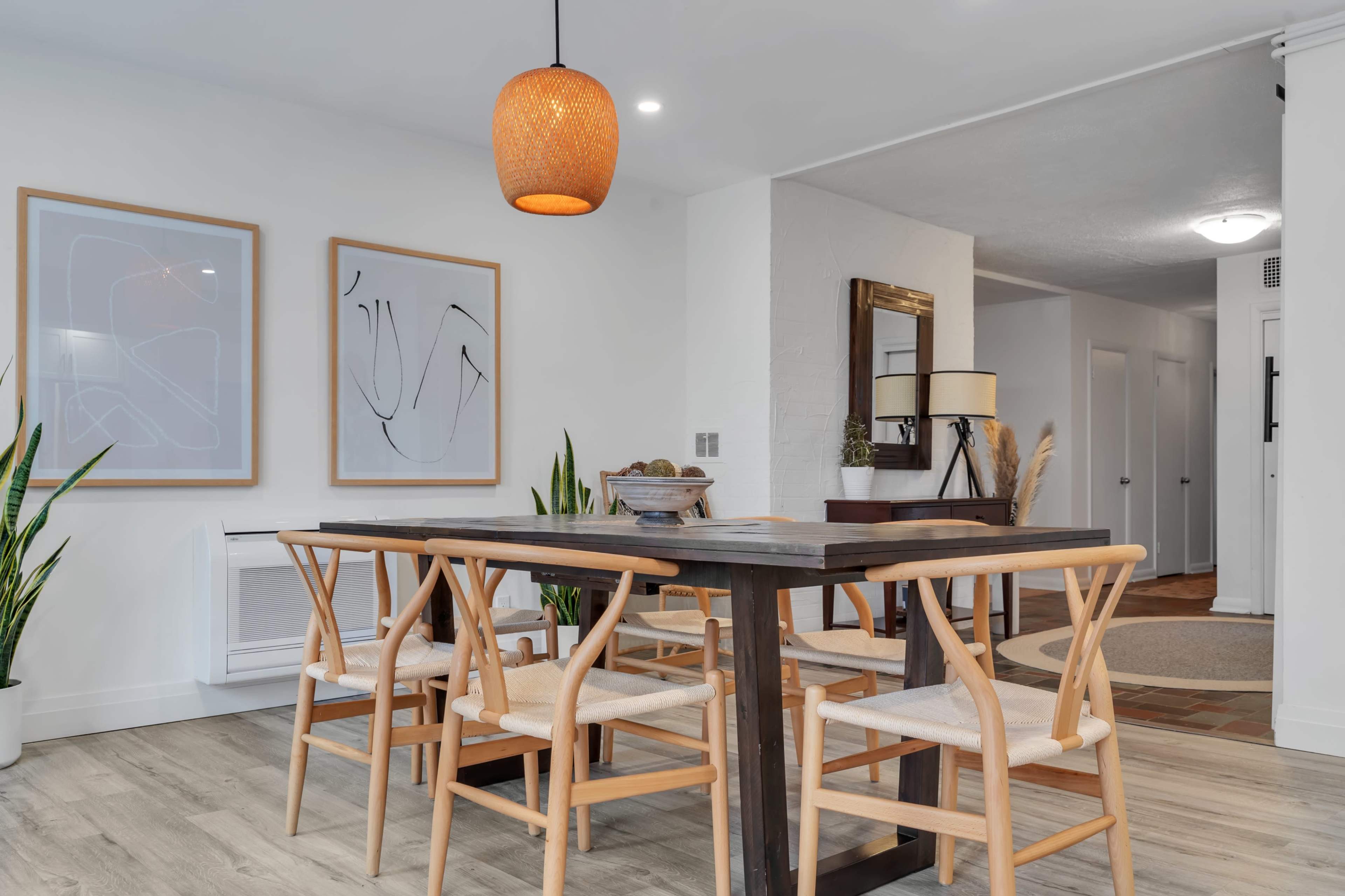 A dining area features a wooden table surrounded by four chairs, with two framed artworks and decorative plants against a white wall.