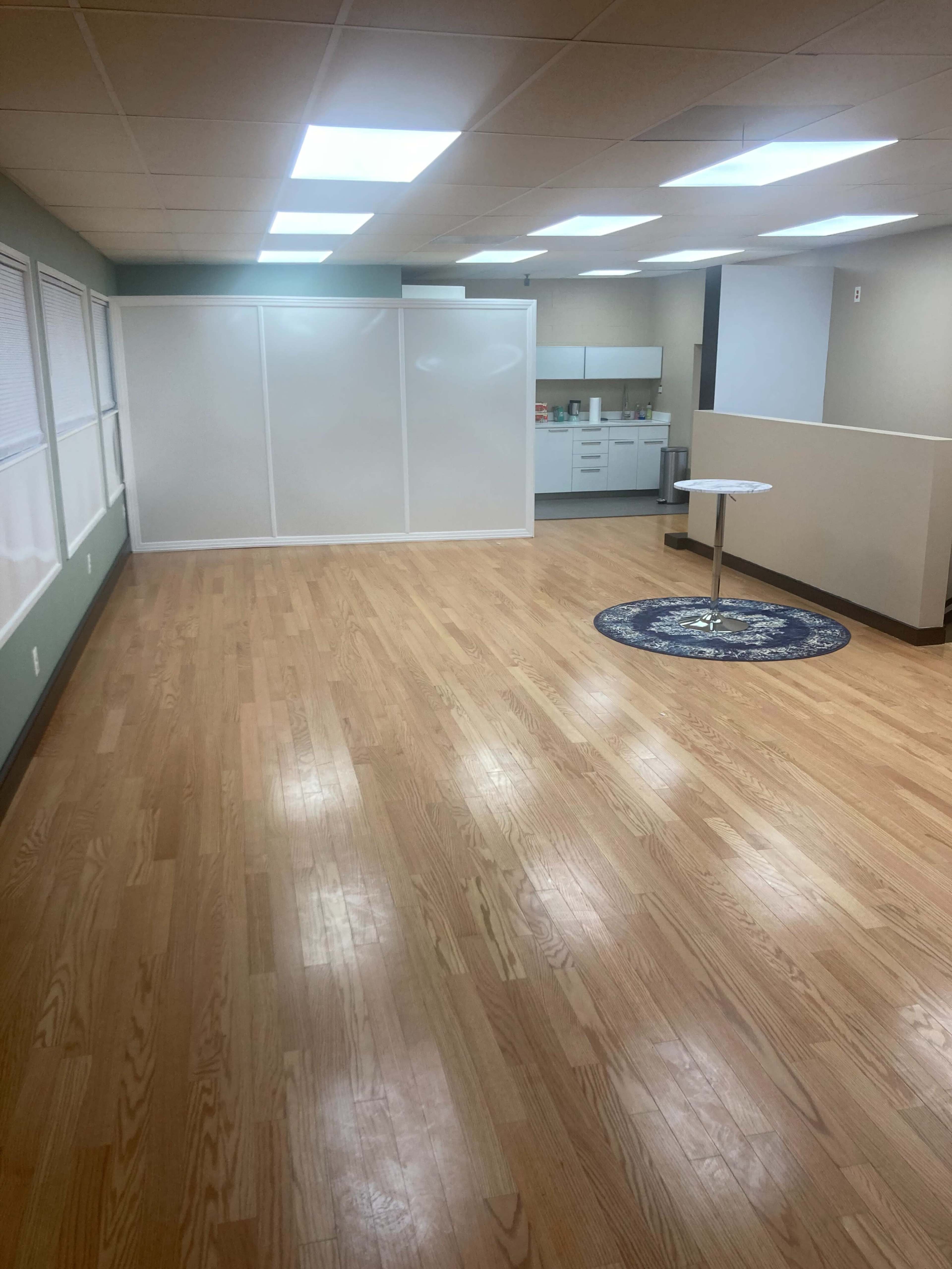 A spacious, empty room with wooden flooring, featuring a small table and a kitchenette area in the background, separated by a partial partition.