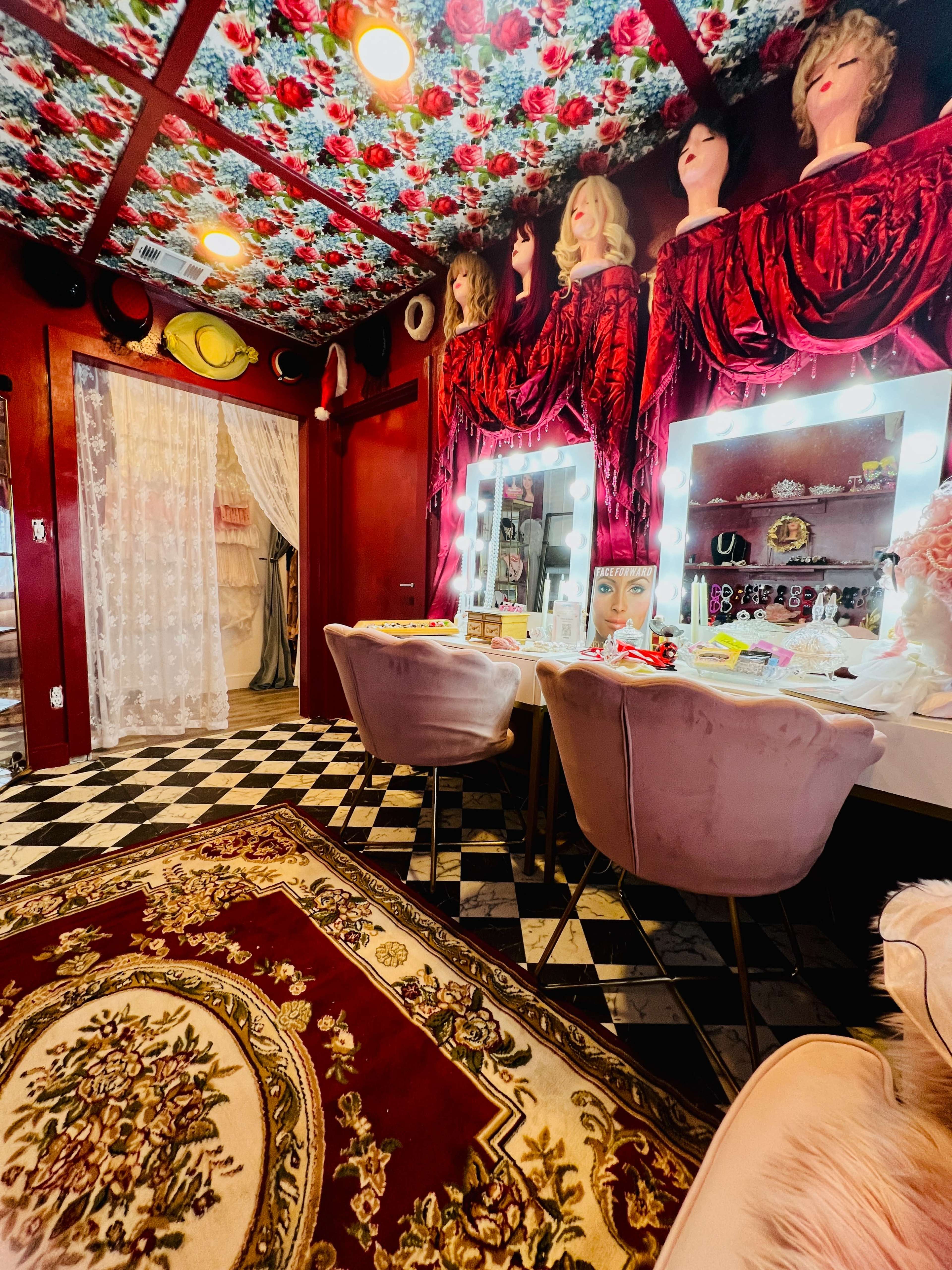 The room features a checkered floor, vintage mannequins with wigs on the ceiling, and vanity mirrors framed by red drapery.