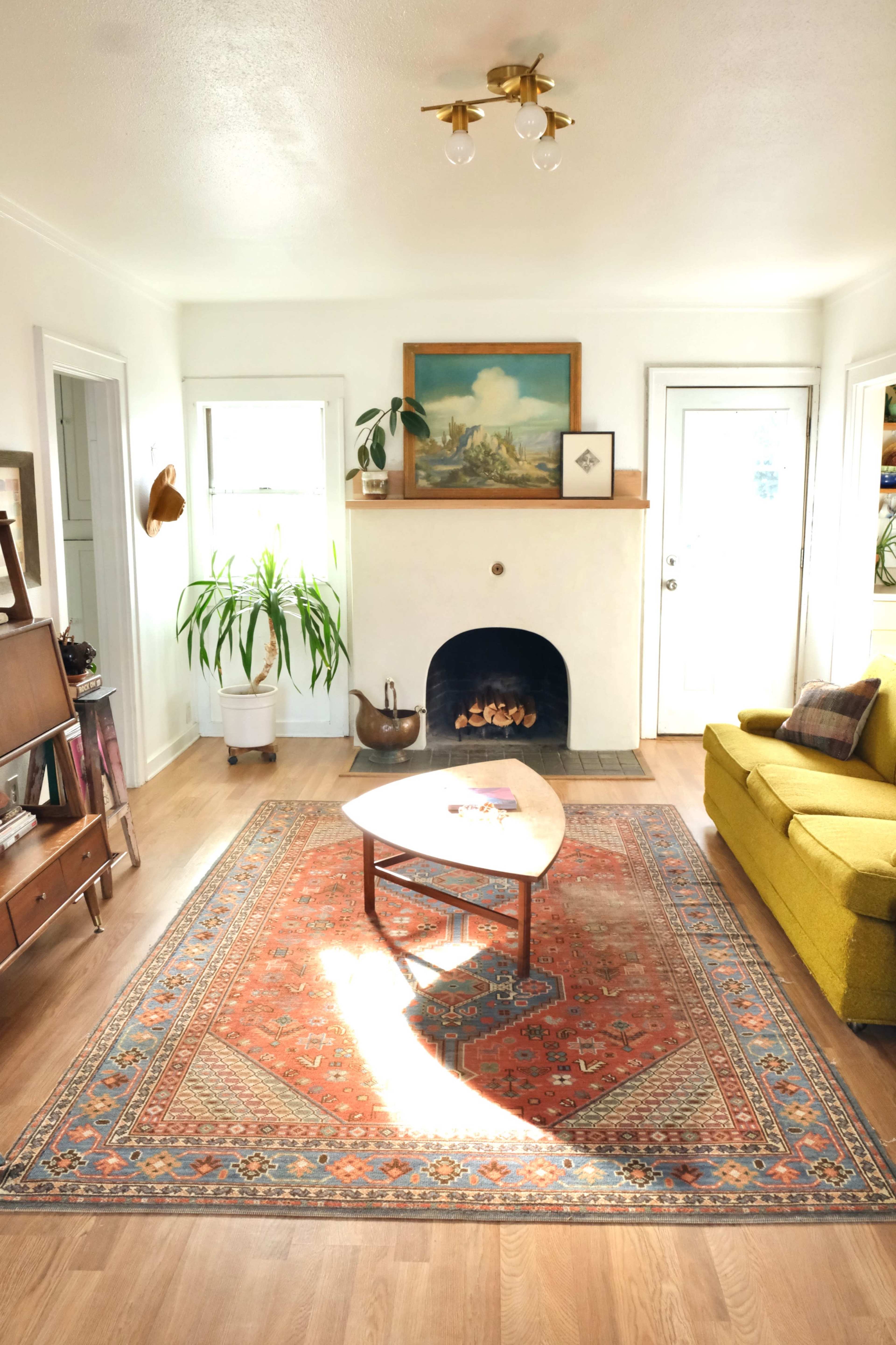 A bright living room features a green sofa, a coffee table on a patterned rug, and a decorative fireplace with a painting above it.