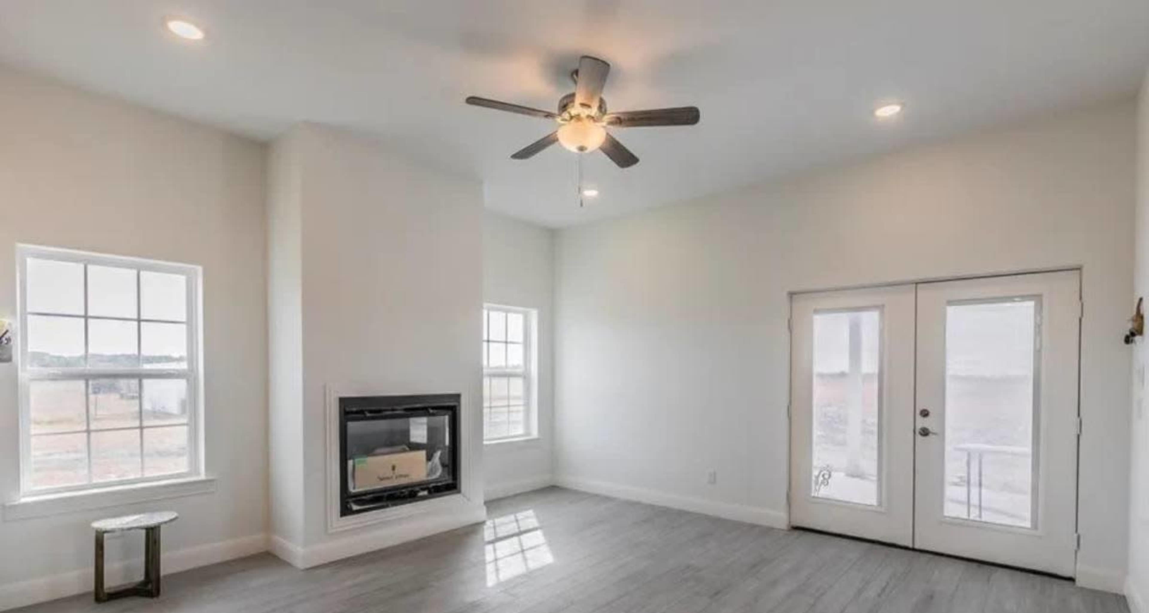The image shows a light-filled living room with a ceiling fan, a fireplace, and large windows, featuring a door that leads outside.