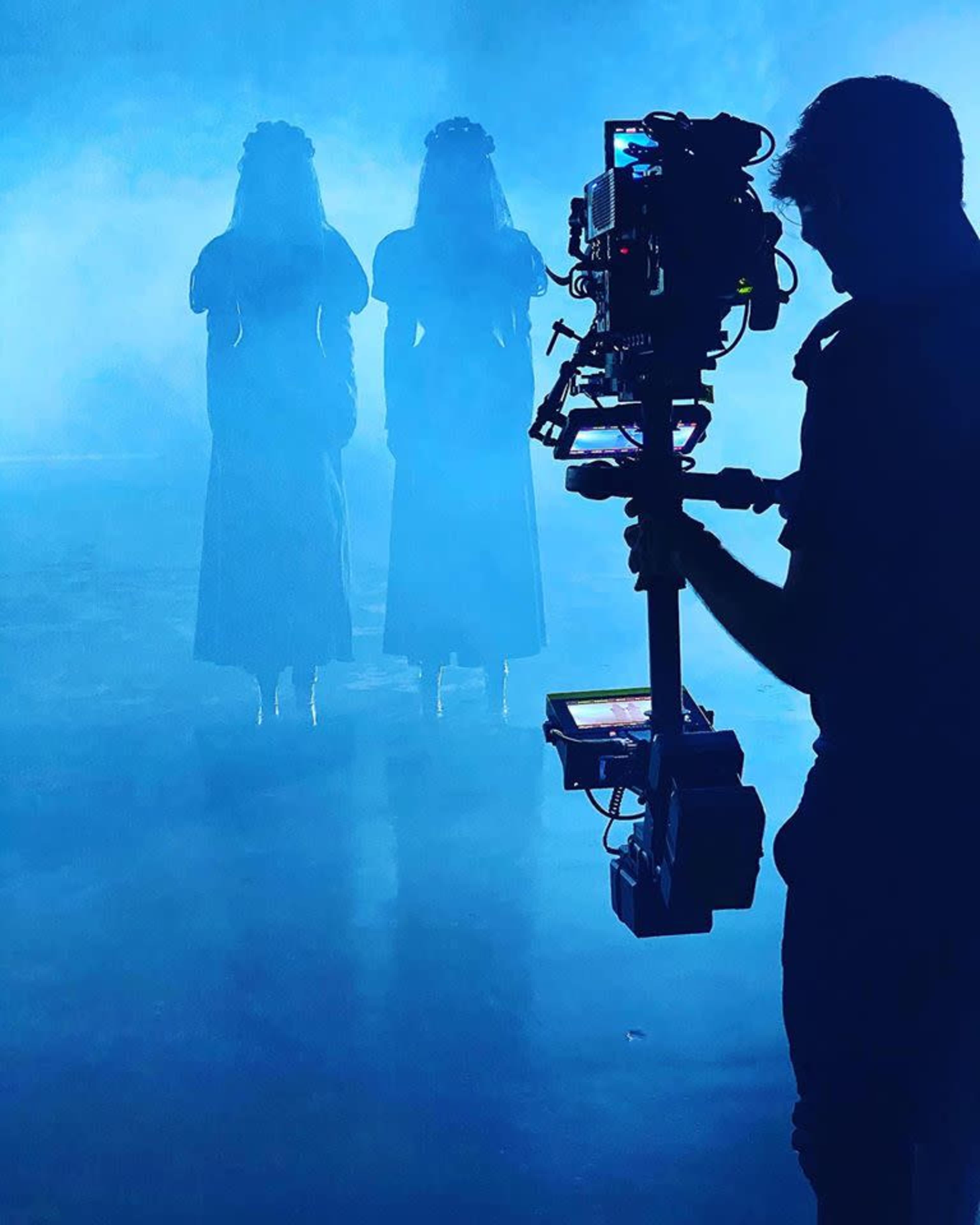 A camera operator captures two figures dressed in long dresses, standing in a foggy, blue-lit environment.