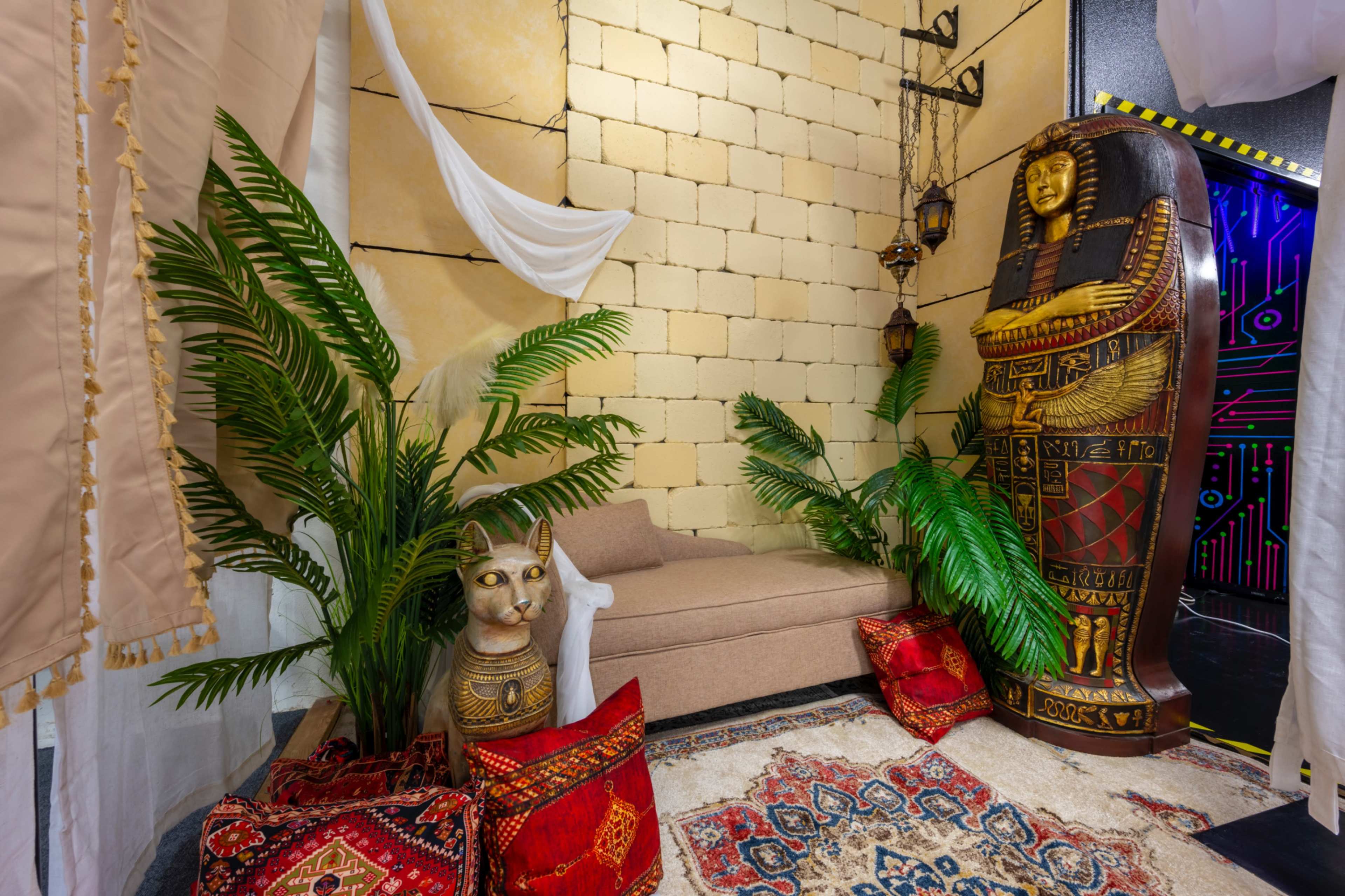 The scene features a cozy corner adorned with a large golden statue of a pharaoh, a cat figure, decorative cushions, and lush greenery against a backdrop of textured wall panels.