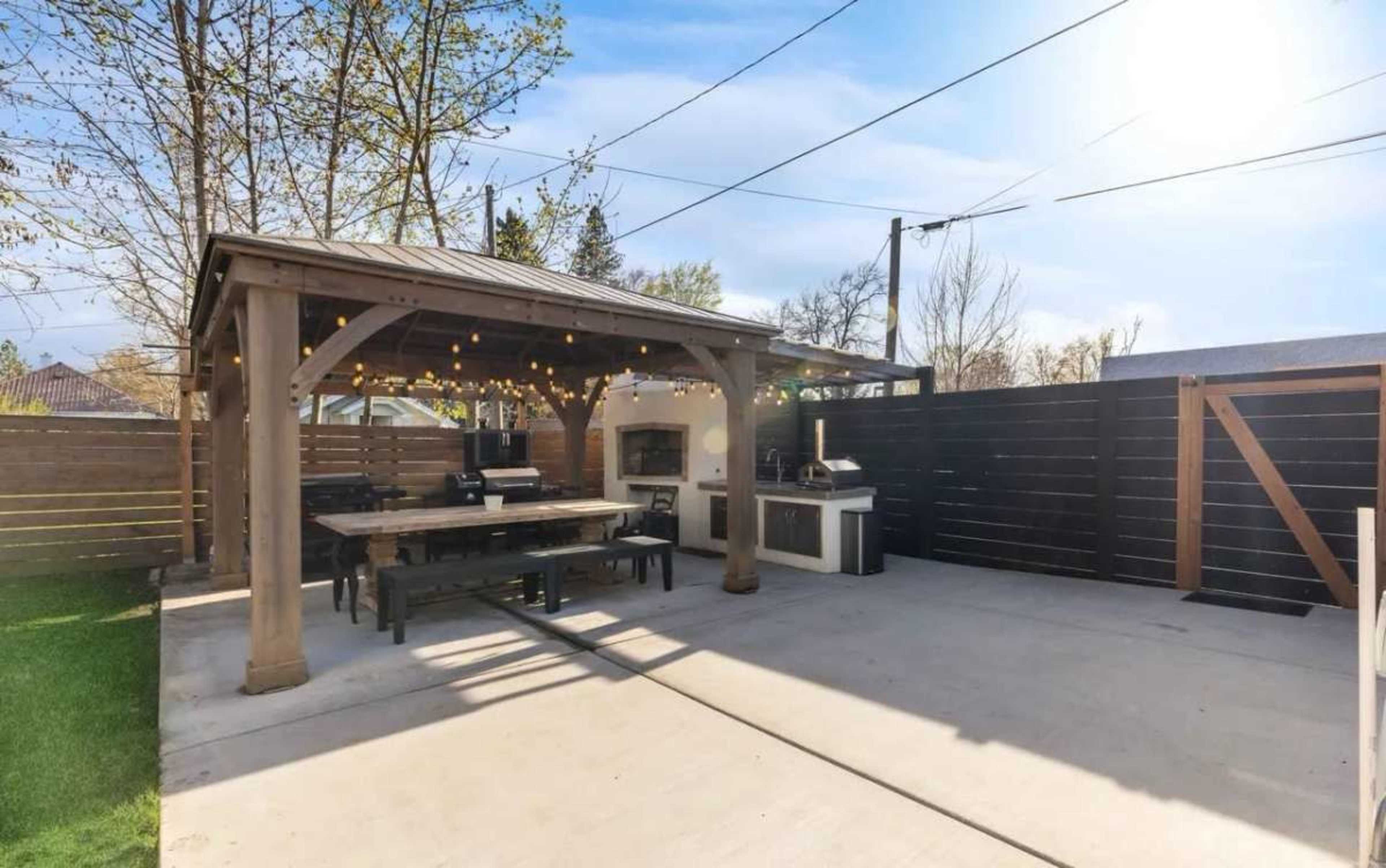 The image shows a spacious outdoor patio area featuring a wooden gazebo, dining table, and outdoor kitchen with a pizza oven.