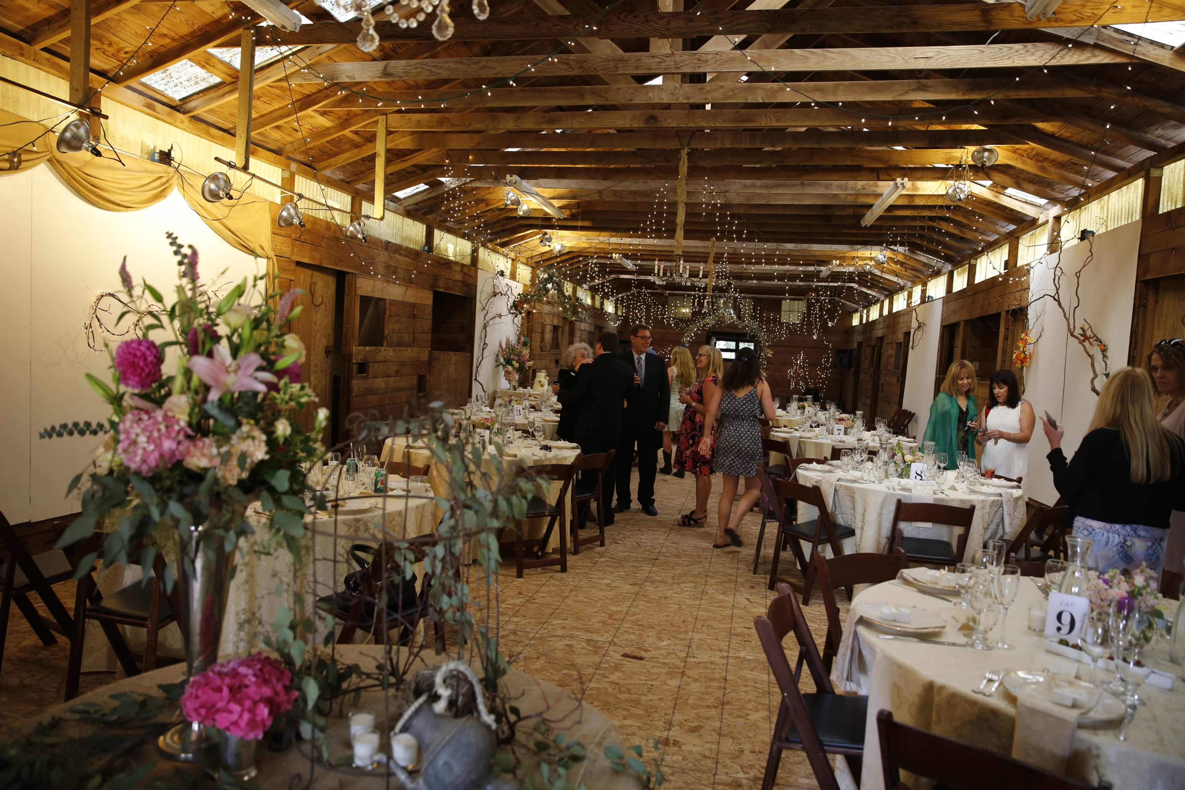 The image shows a decorated barn interior with tables set for a gathering, surrounded by floral arrangements and string lights.