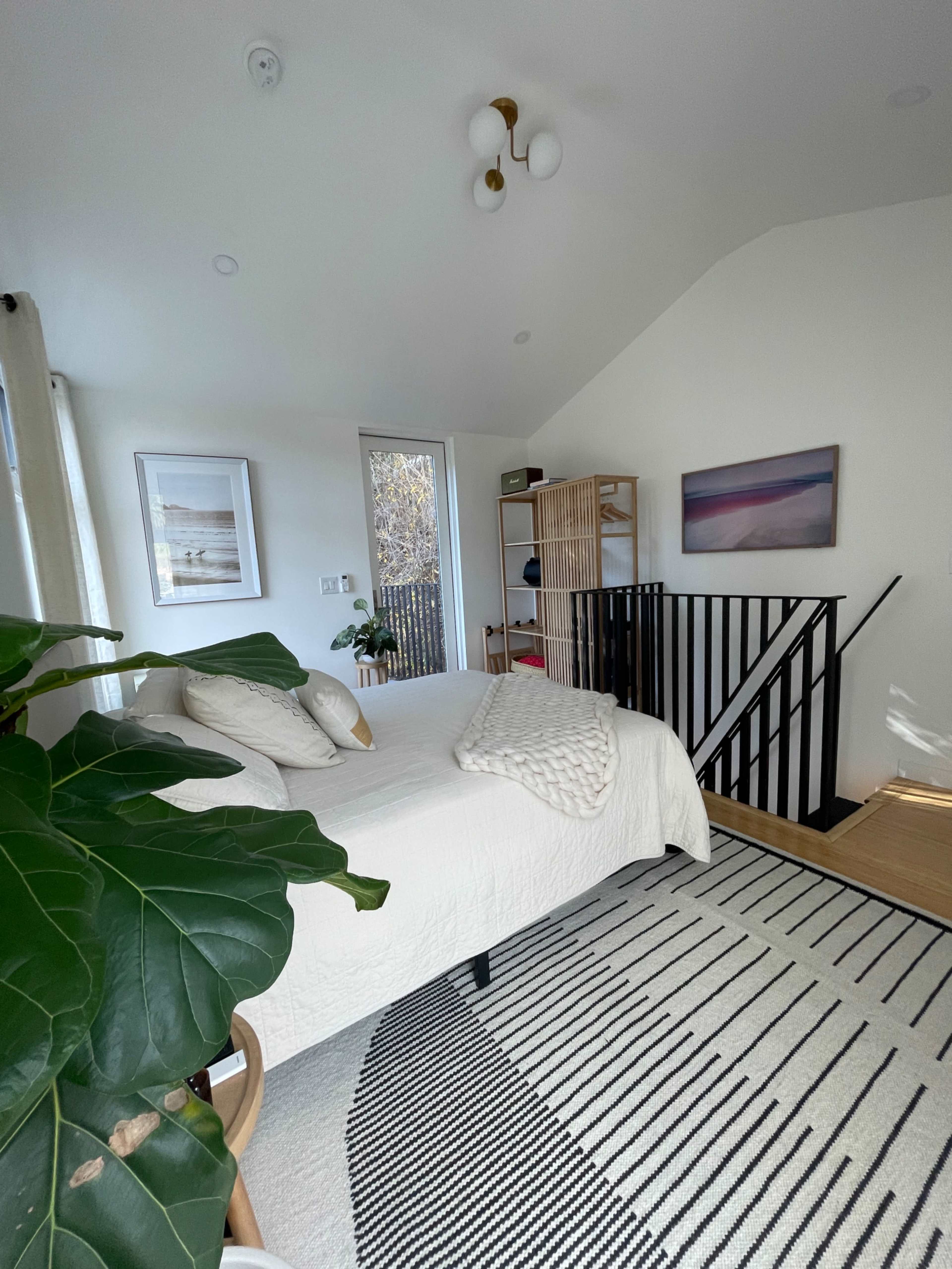 The image shows a bright bedroom with a bed, a patterned rug, and a staircase leading to another area.