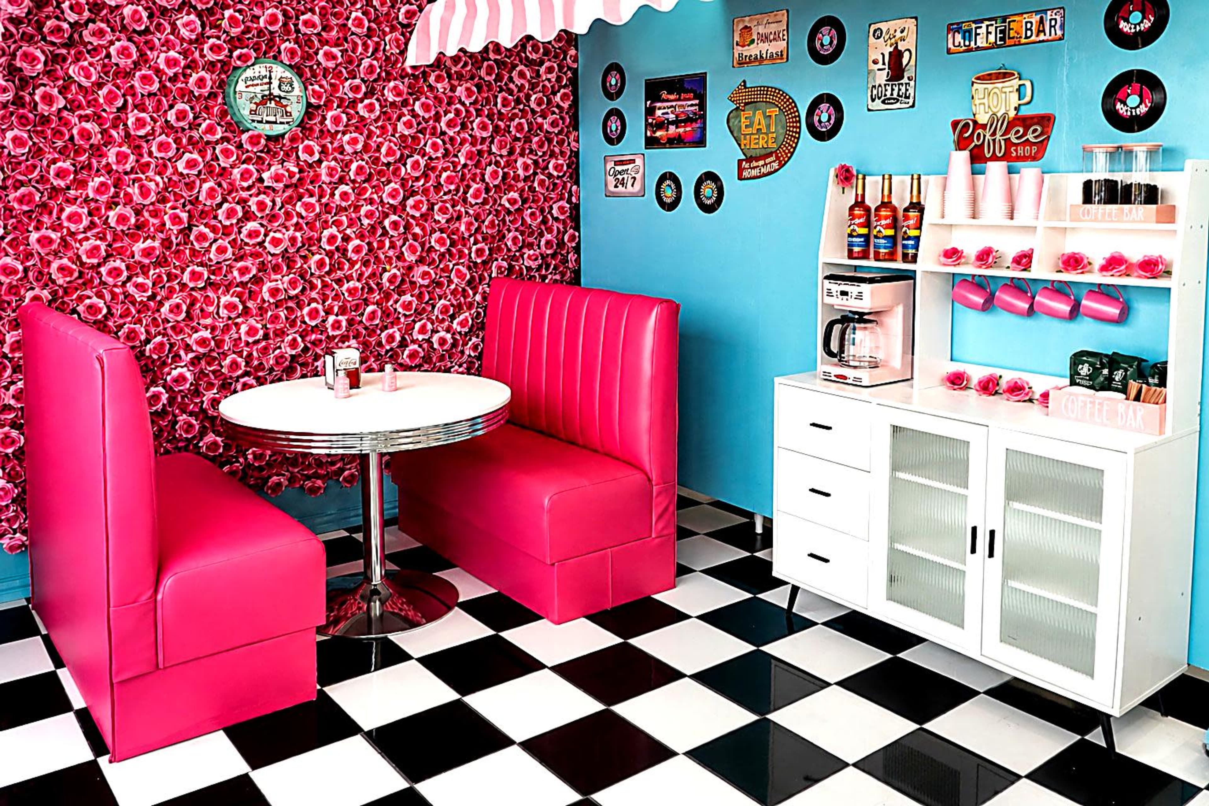 The image shows a colorful diner interior with pink booths, a round white table, a patterned backdrop, and a coffee station against a blue wall.