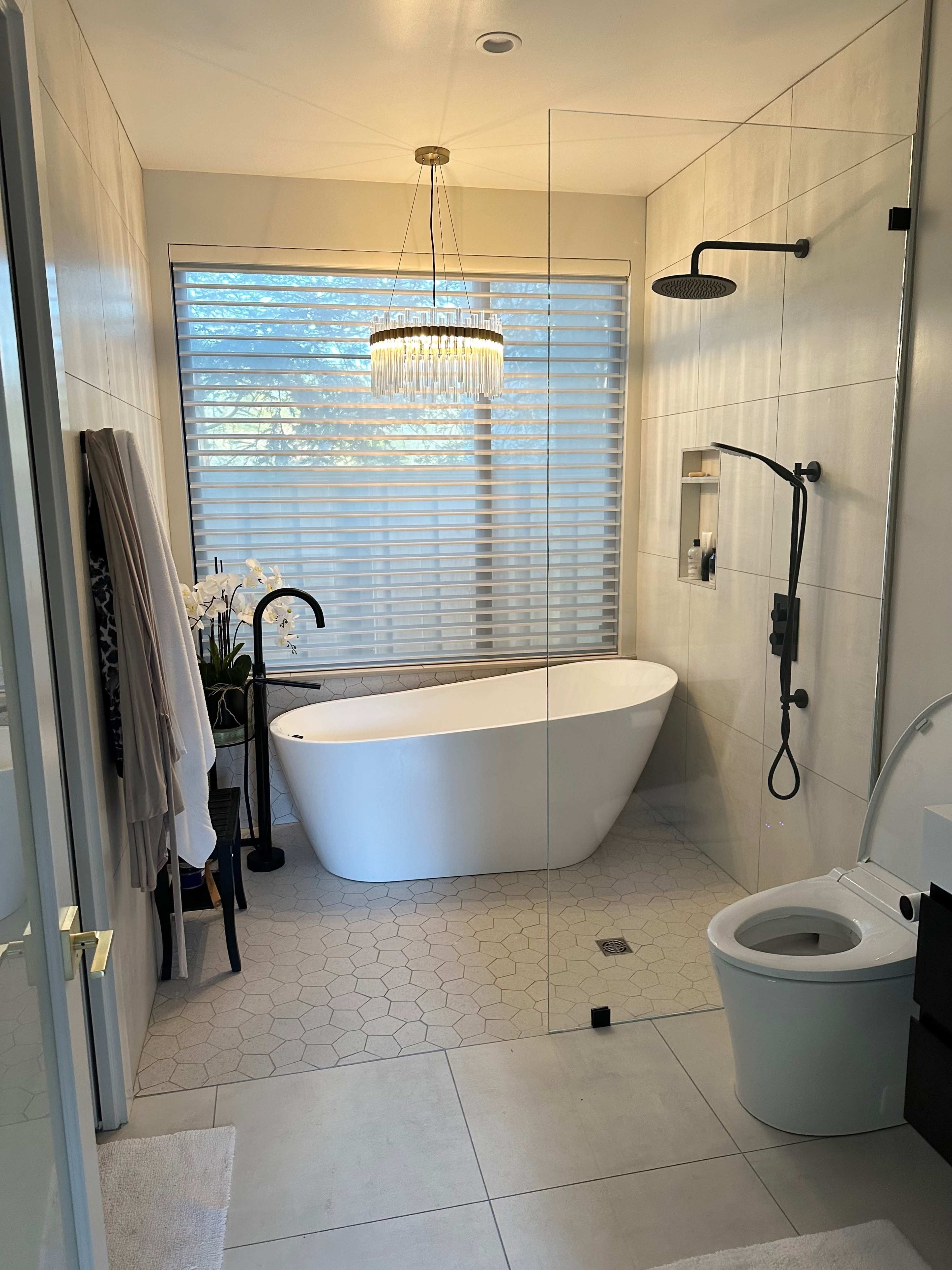The bathroom features a freestanding tub, a glass shower enclosure, a modern toilet, and a stylish chandelier above, with large windows allowing natural light to filter through the blinds.