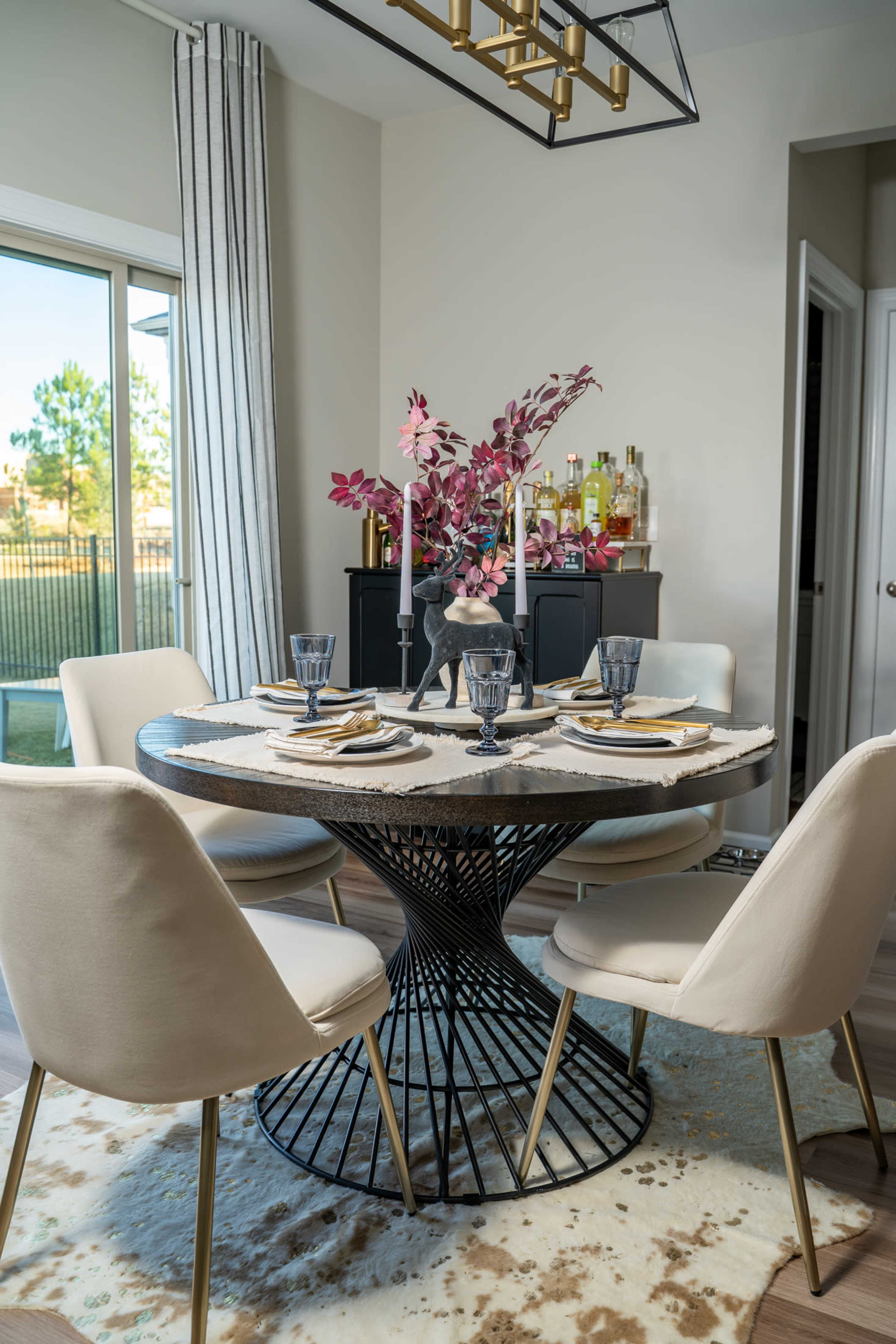A round dining table is set with plates and glasses, surrounded by four modern chairs, in a well-lit room with a view of an outdoor area.
