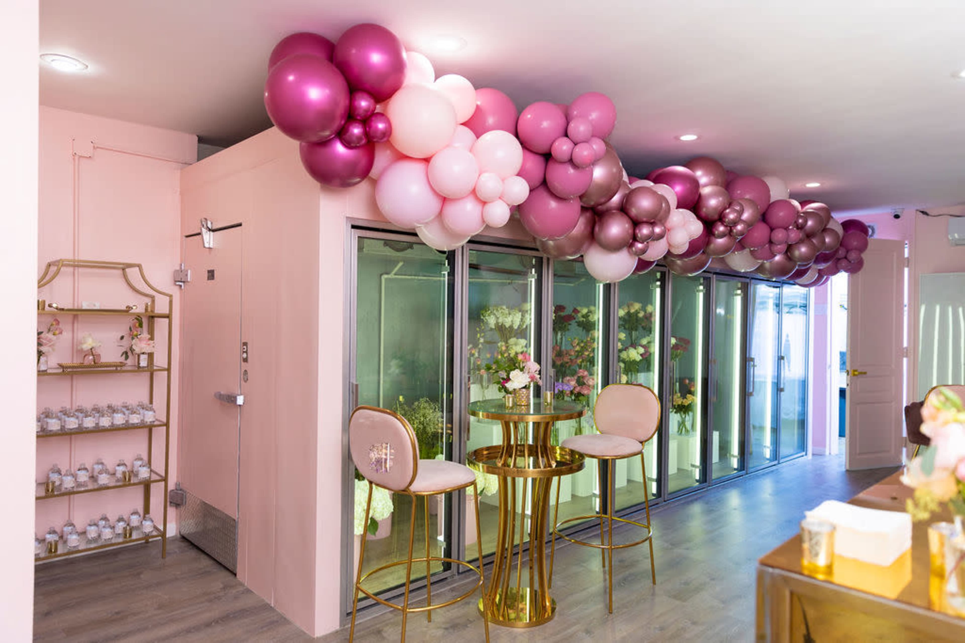 The image shows a decorated room with a balloon arch in shades of pink and purple, a small round table with two stools, and a shelf displaying small containers.