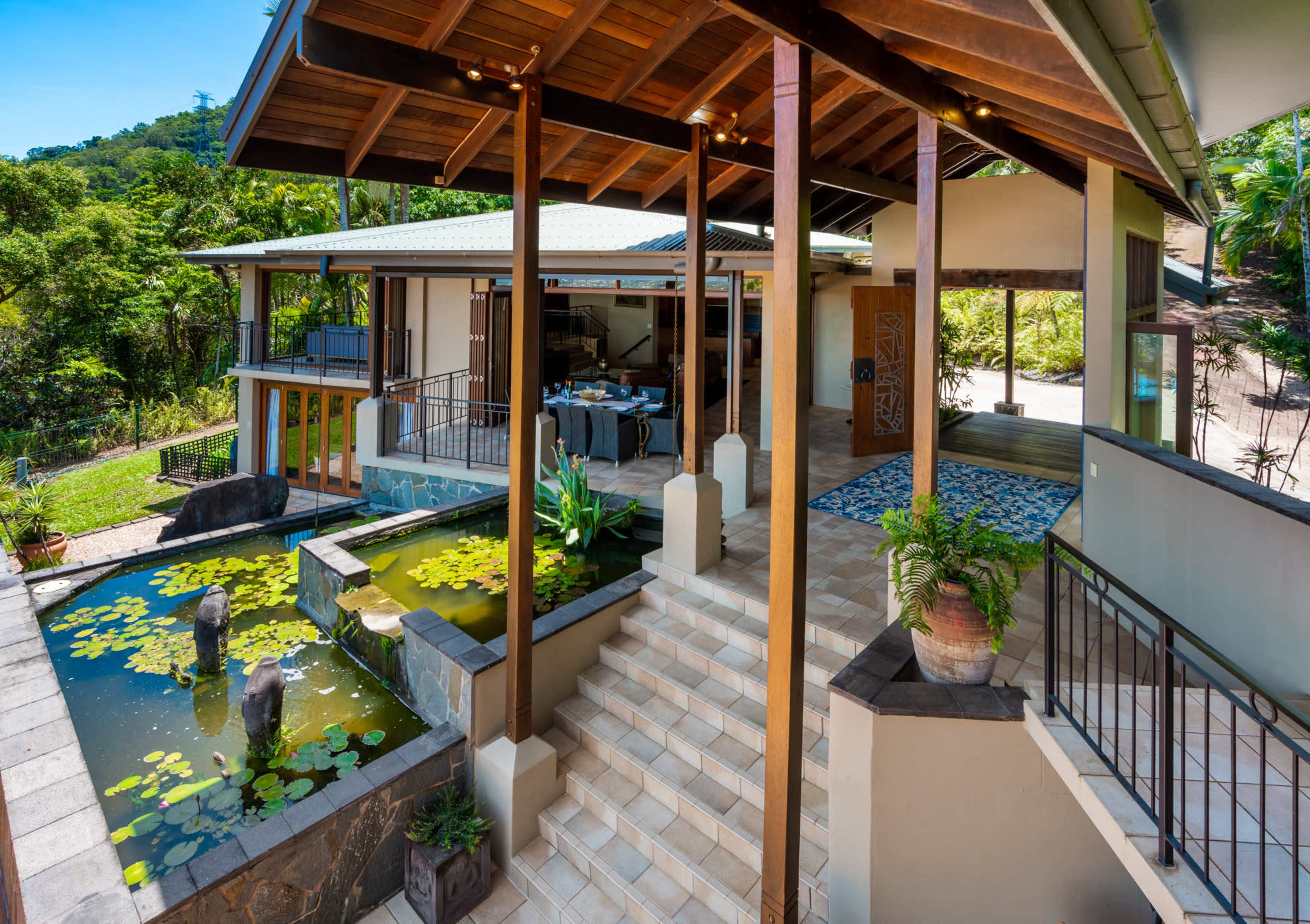 The image shows a modern house with an open design featuring a pond surrounded by greenery, stairs leading to the entrance, and a dining area visible inside.
