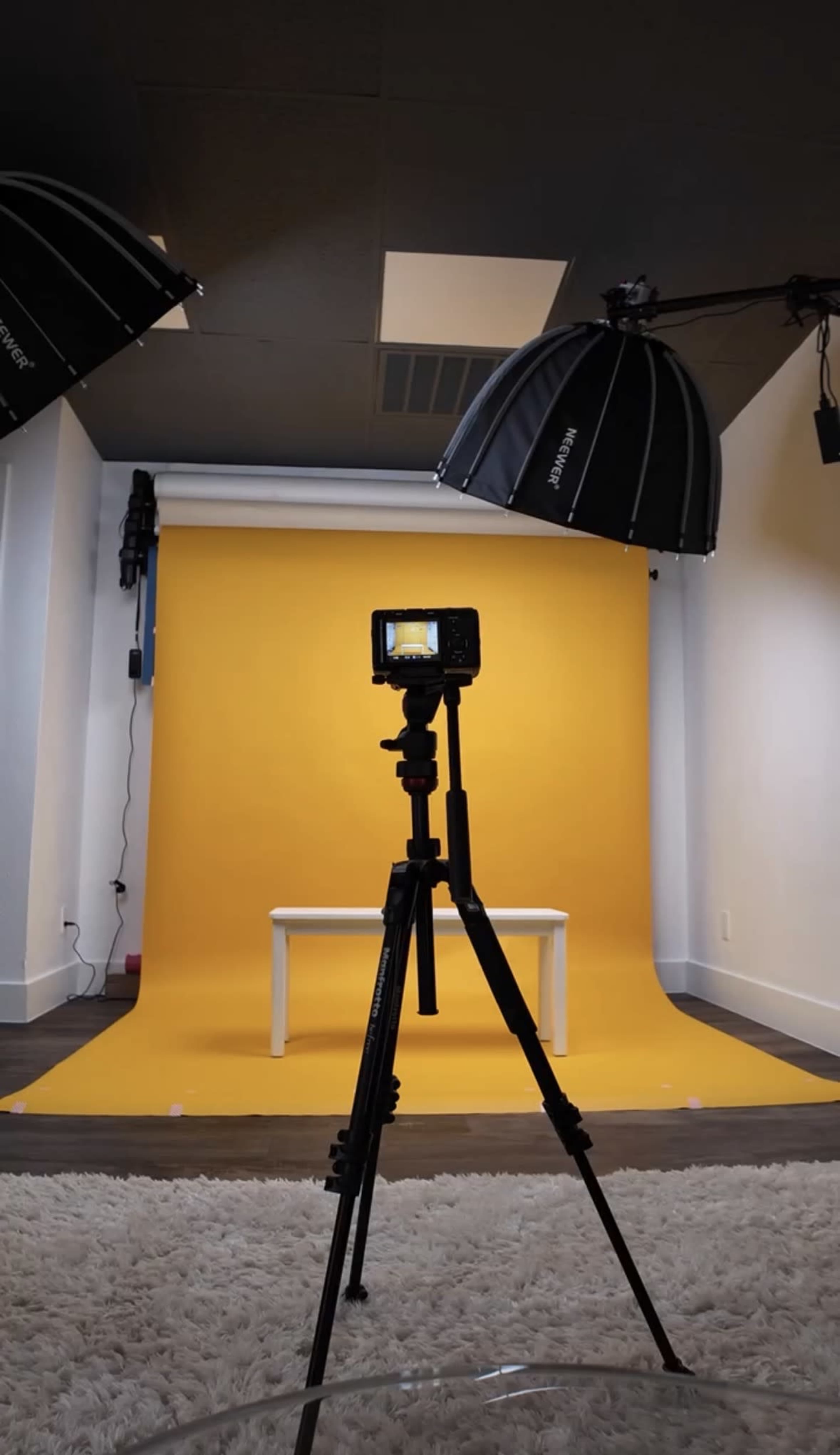 The image shows a photography studio setup with a yellow backdrop, a white table in front of it, and softbox lights positioned above.