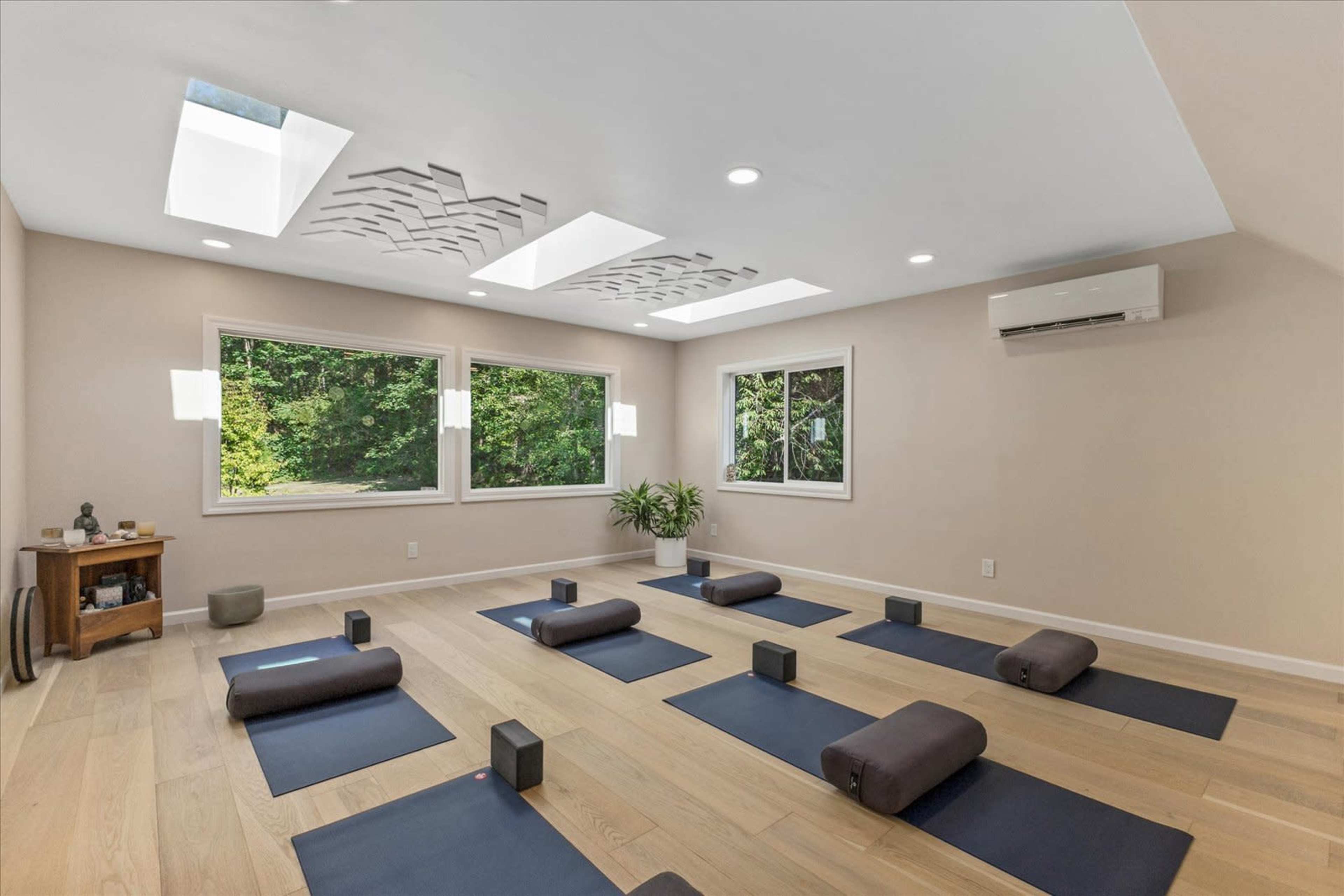 The image shows a serene yoga studio with mats, blocks, and bolsters arranged in neat rows beneath skylights and large windows that allow natural light to enter.