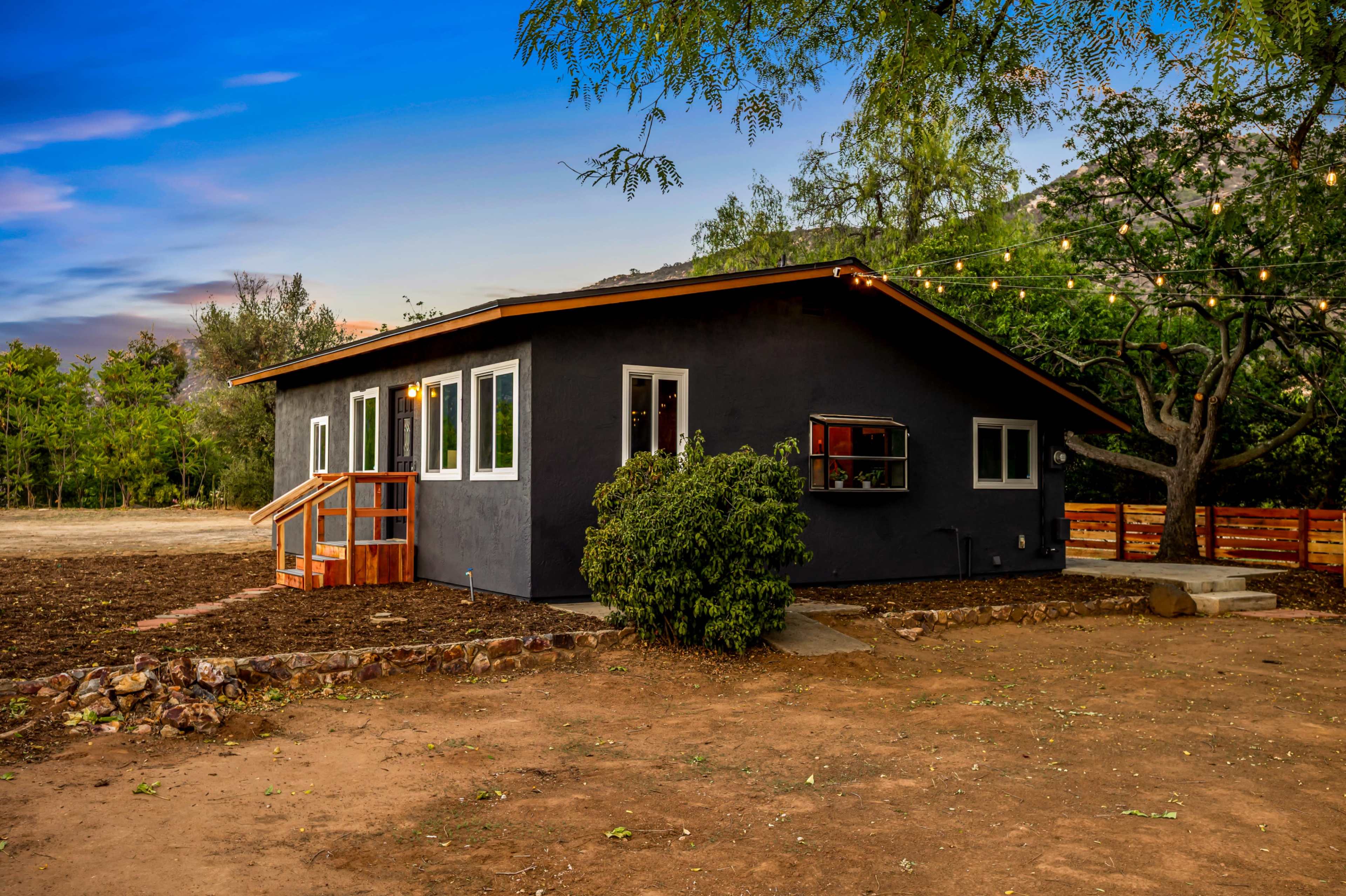 A dark-colored house with multiple windows is surrounded by a cleared area and a few trees, with outdoor string lights visible above.