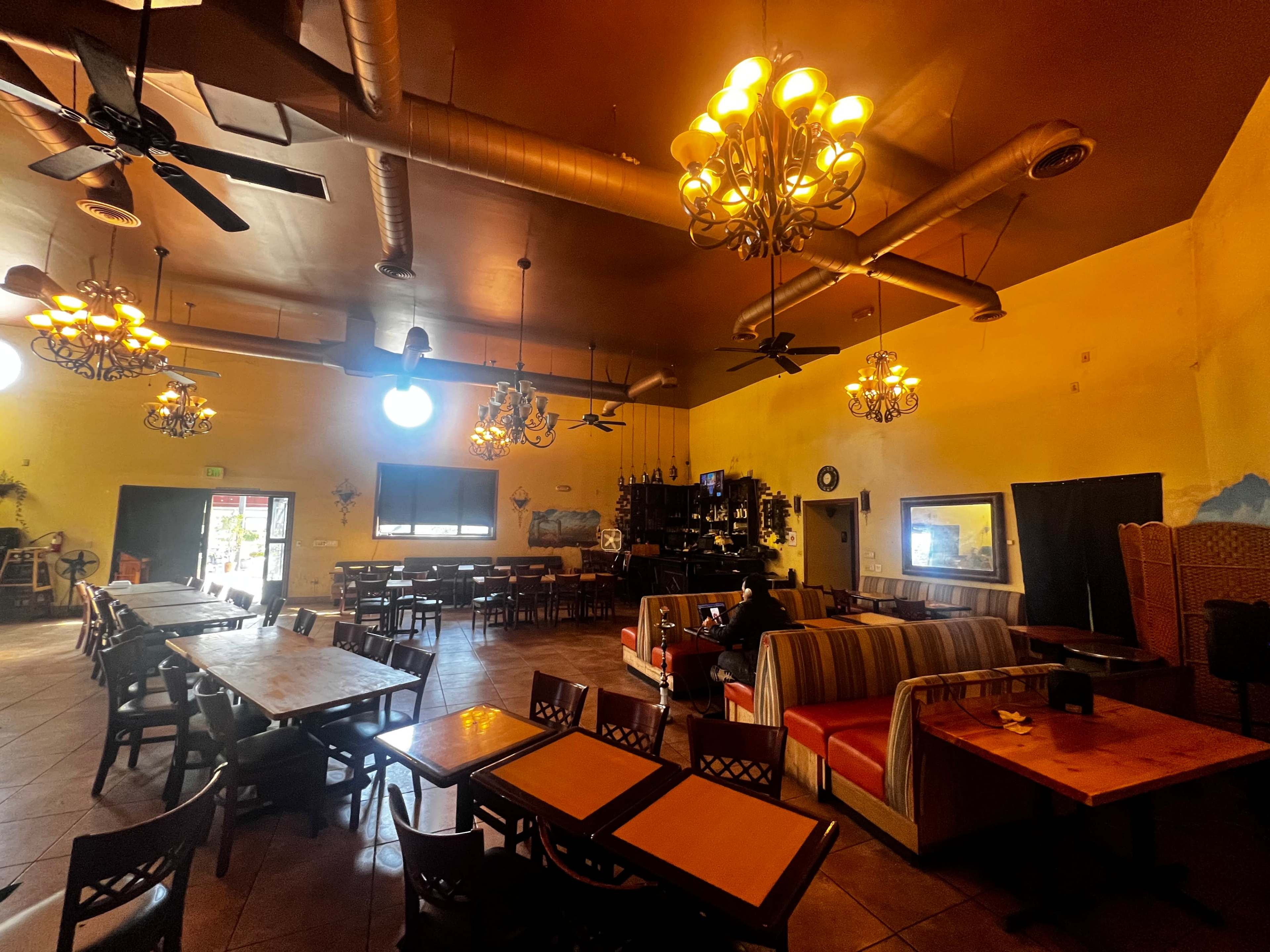 A spacious restaurant interior with wooden tables, chairs, and ceiling fans, illuminated by several chandeliers.