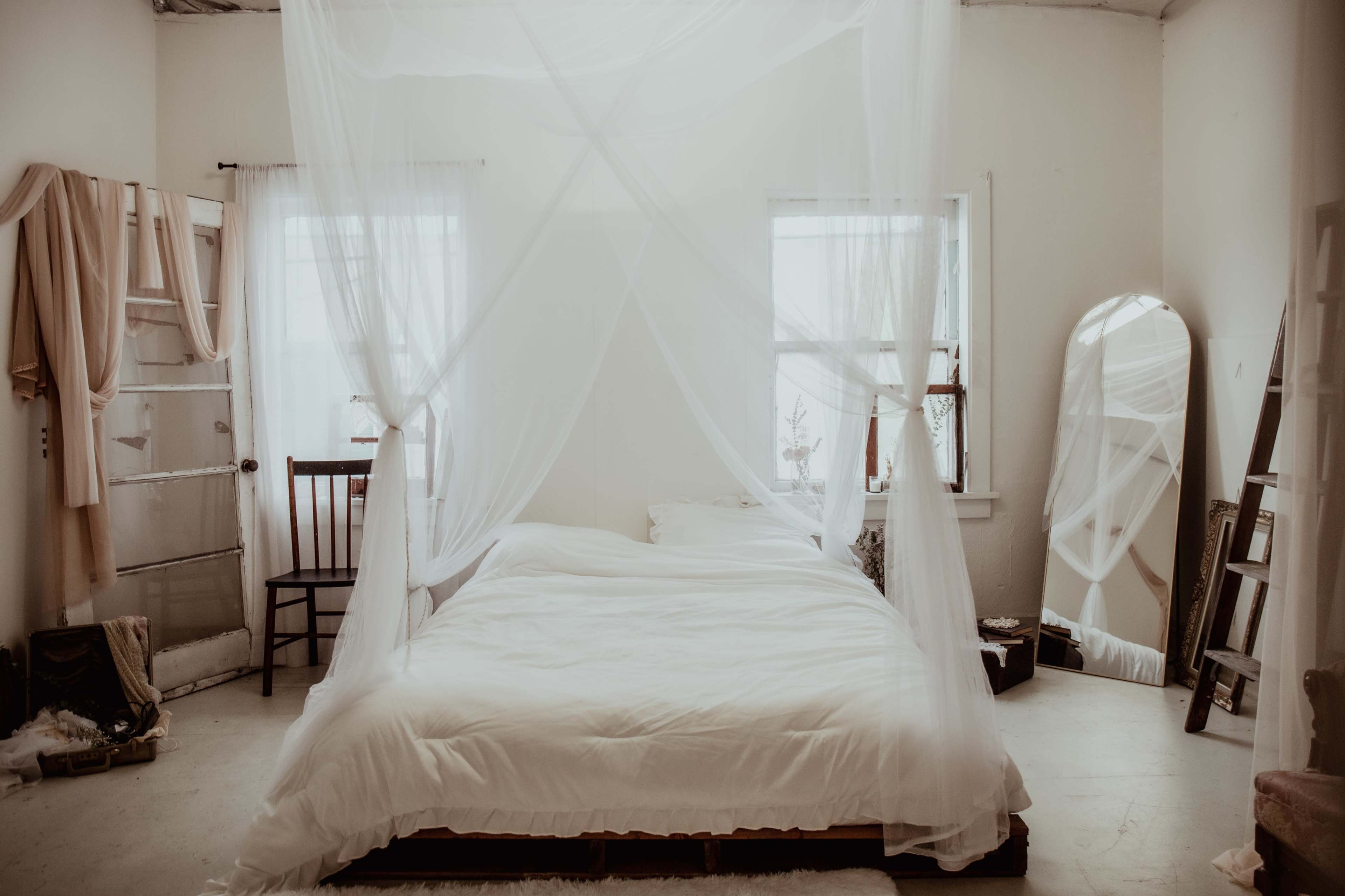 A minimalist bedroom with a large bed draped in sheer white fabric, two windows, and a wooden chair.