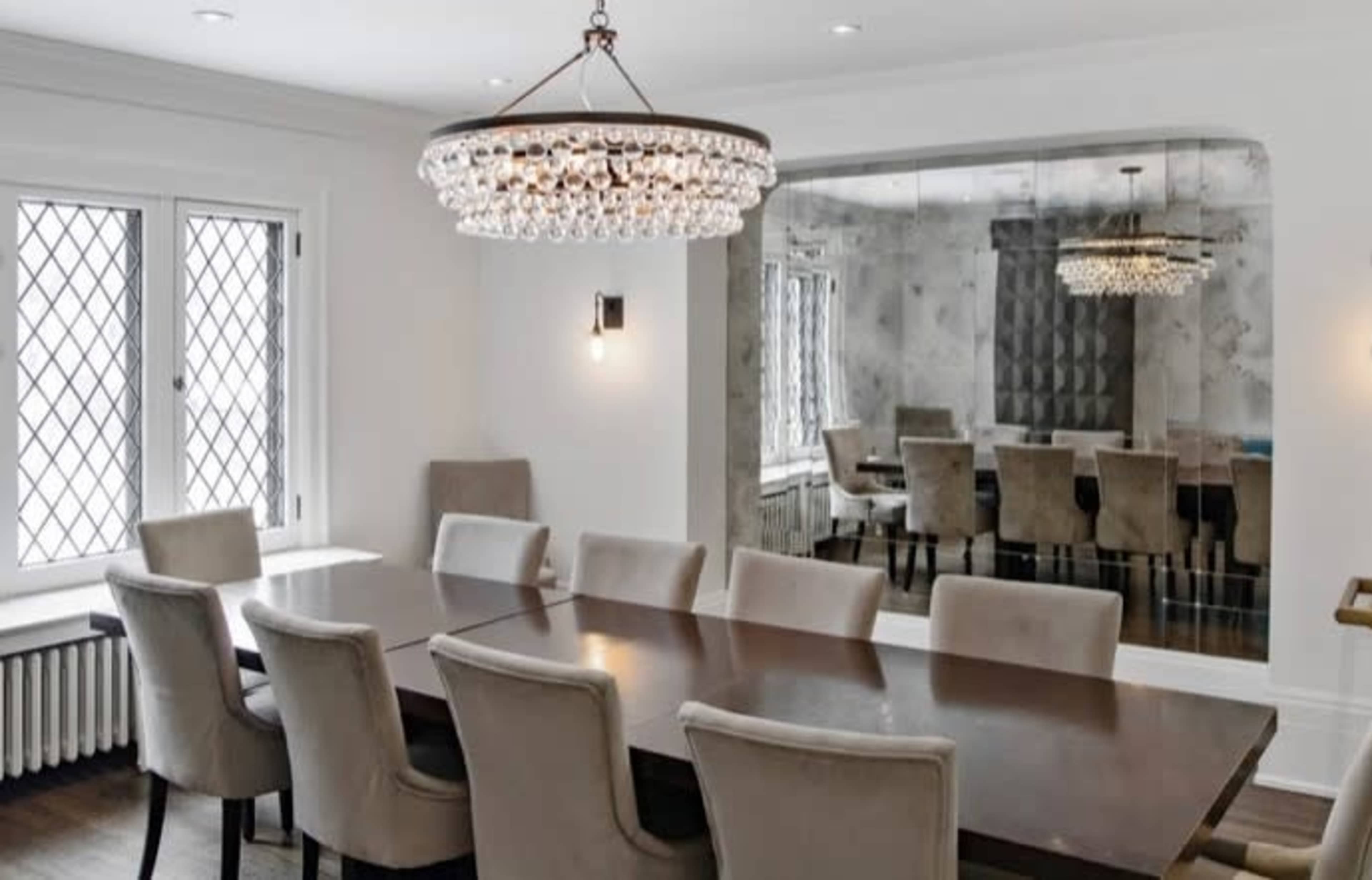 A spacious dining room with a large wooden table surrounded by upholstered chairs, featuring a chandelier and a mirrored wall.