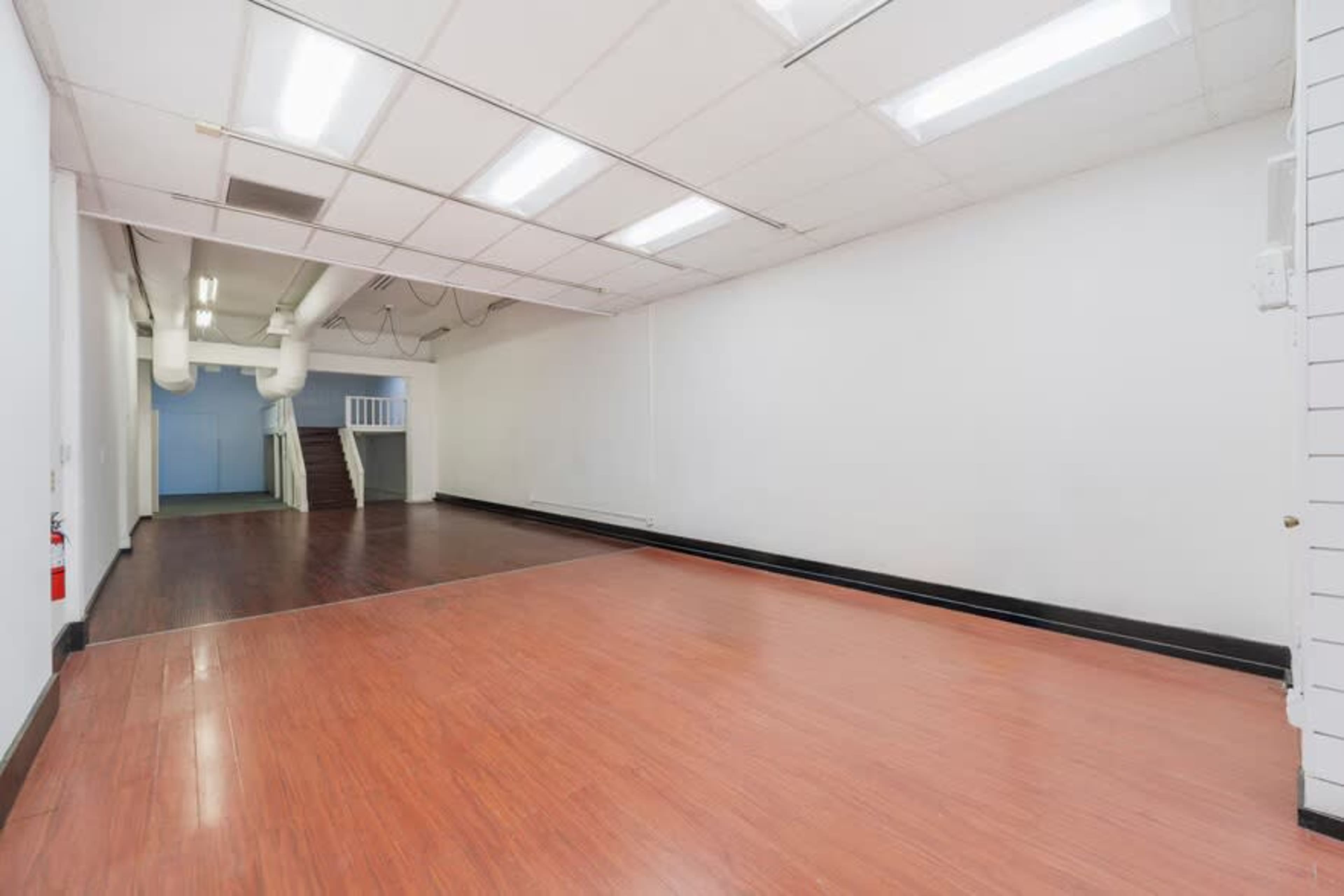 The image shows a large, empty room with a wooden floor and white walls, featuring a double staircase leading to an upper level in the background.