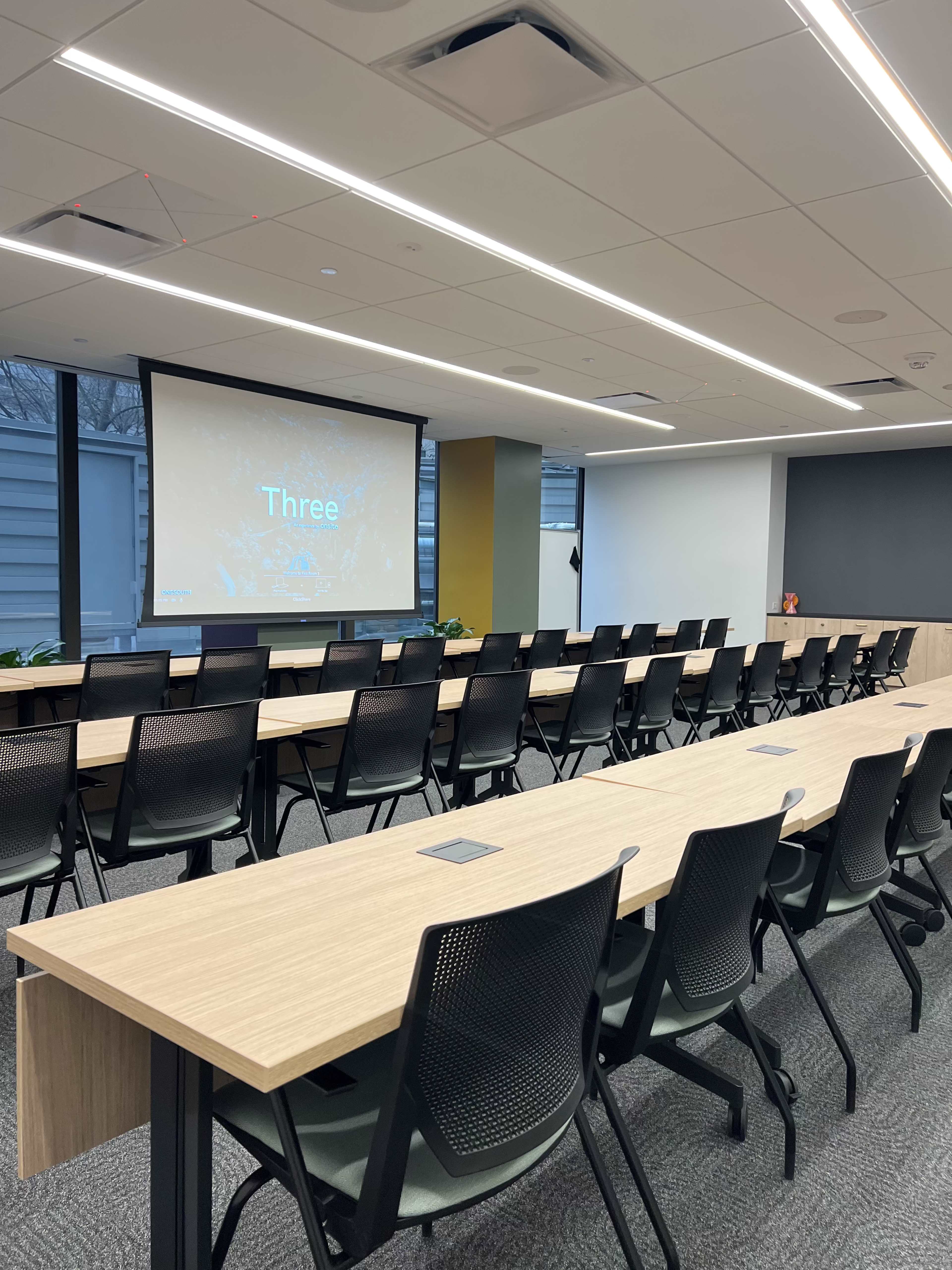 A conference room is arranged with rows of chairs facing a large screen displaying the word "Three."