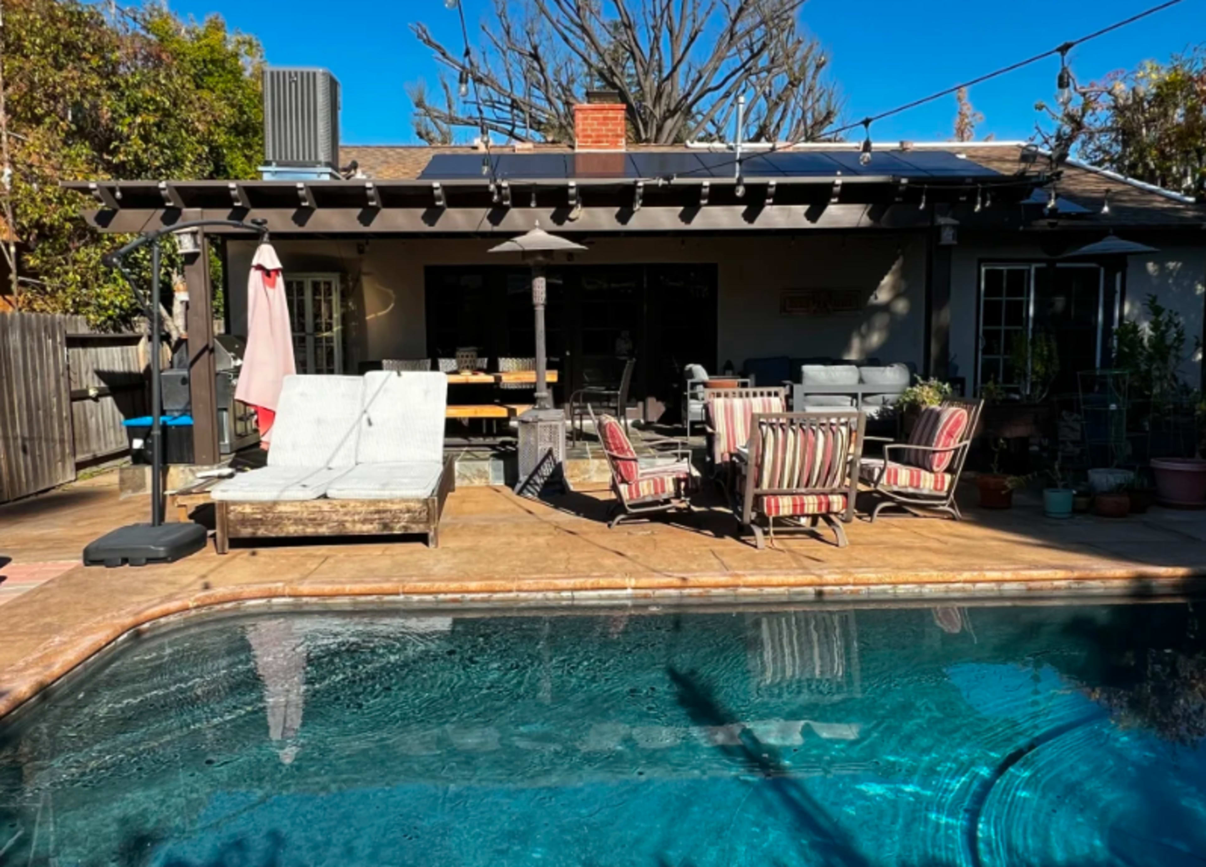 A backyard scene features a swimming pool adjacent to a patio with lounge chairs, dining tables, and shaded areas under a pergola.