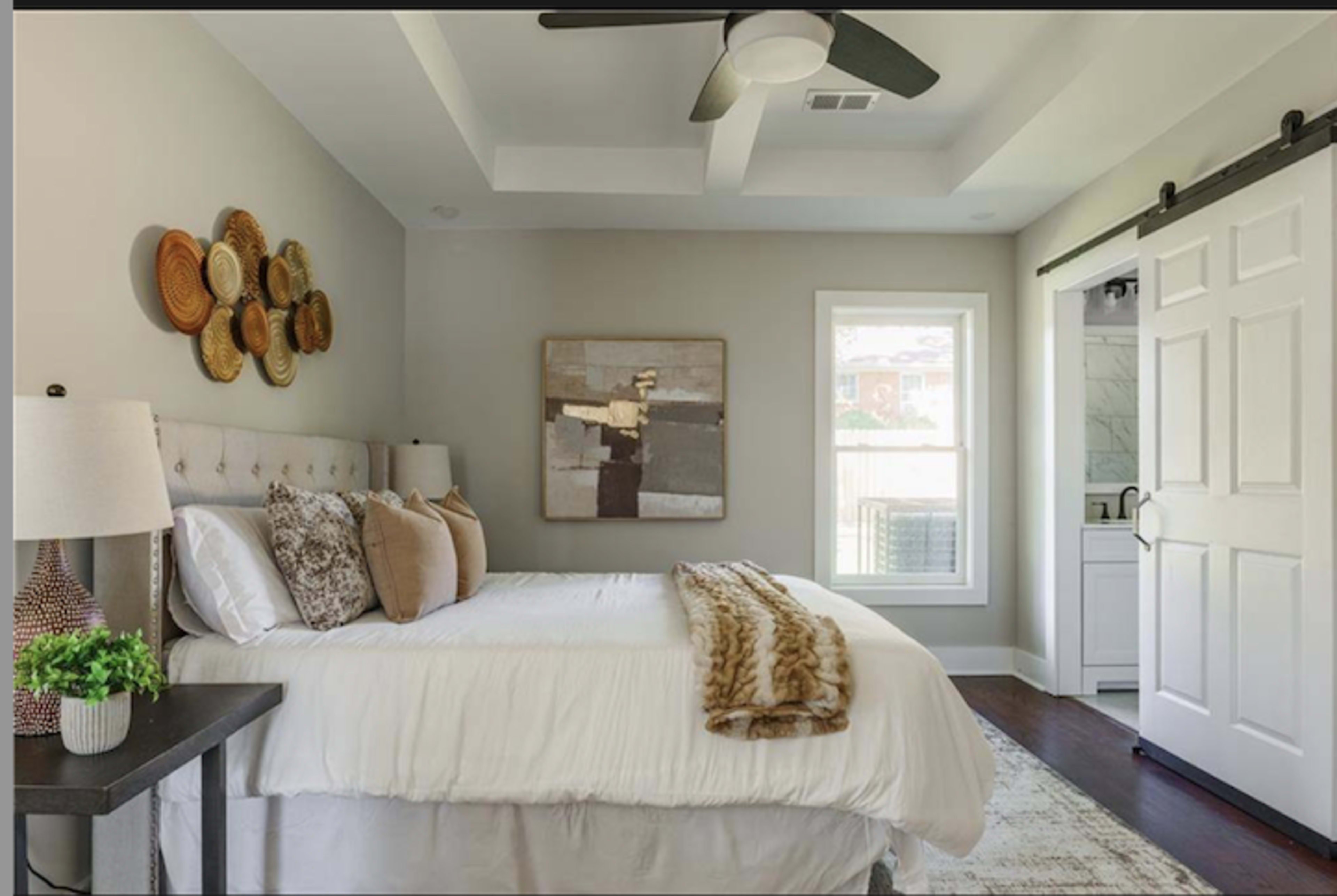 The image shows a well-decorated bedroom featuring a queen-sized bed with decorative pillows, a nightstand with lamps, and a window that opens to natural light.