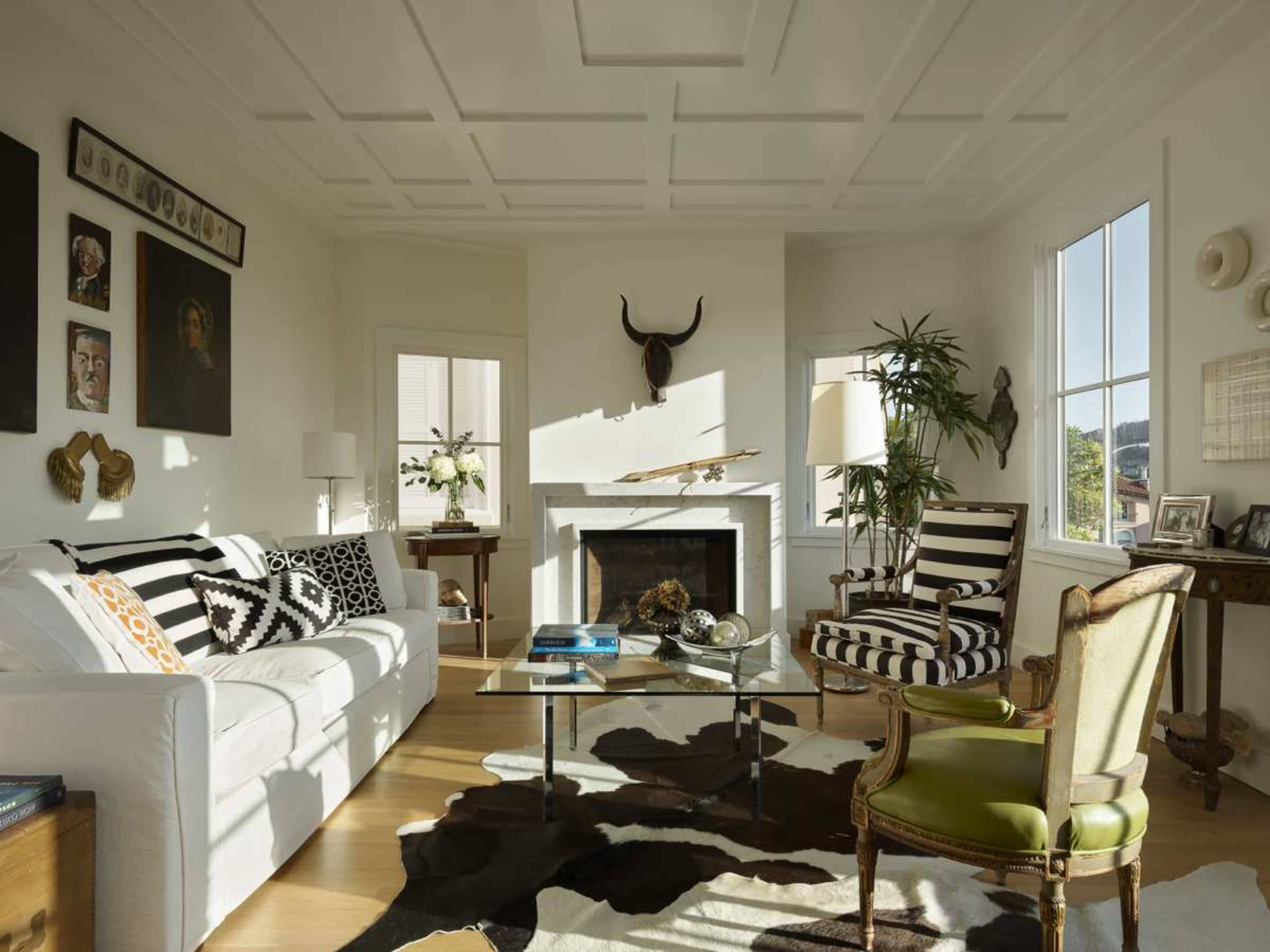 The image shows a modern living room with a white sofa, striped chairs, a glass coffee table, and a cowhide rug, accented by wall art and a decorative fireplace.