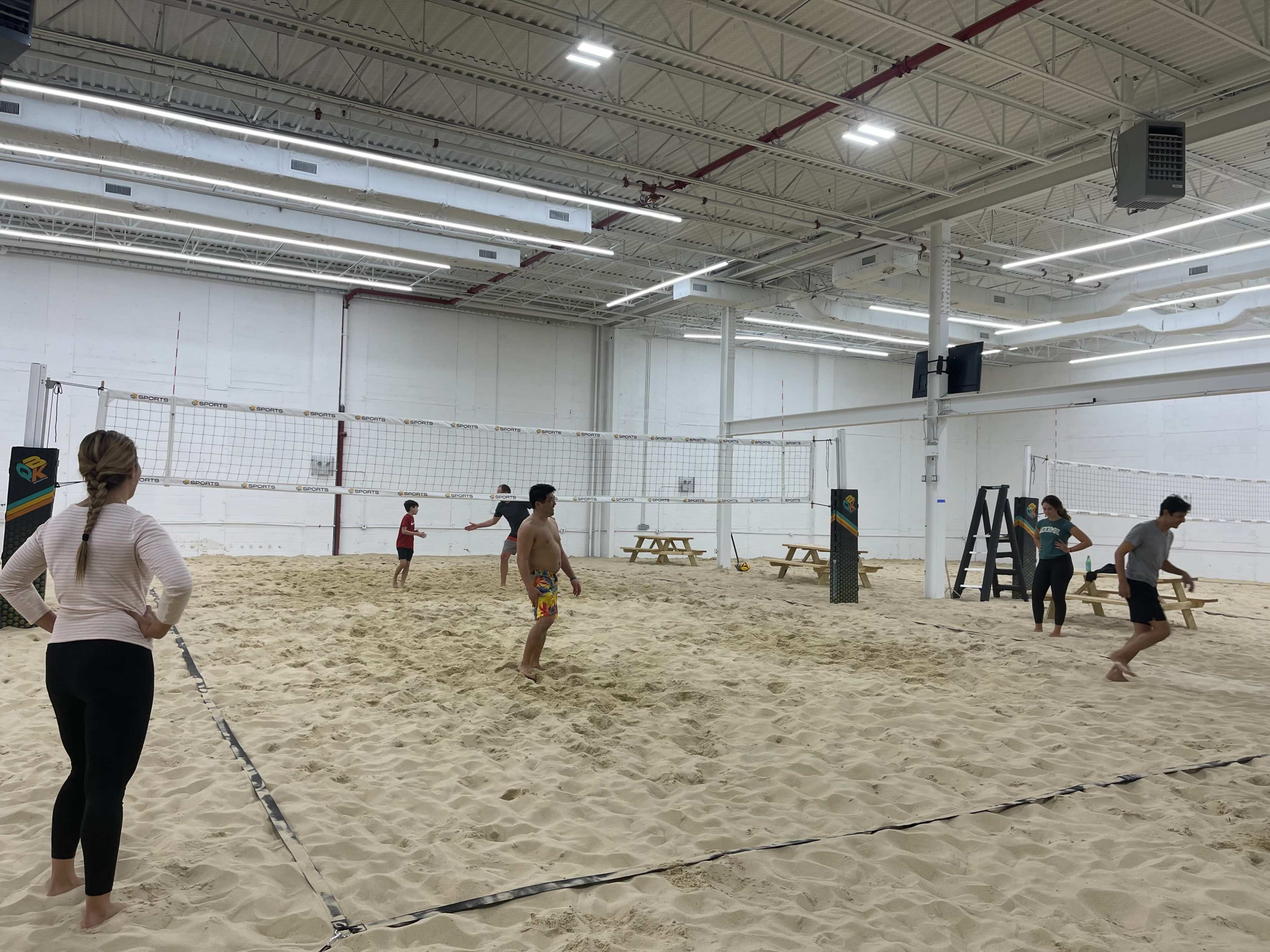 A group of people play volleyball on a sand court indoors, surrounded by a white-walled gym with picnic tables nearby.