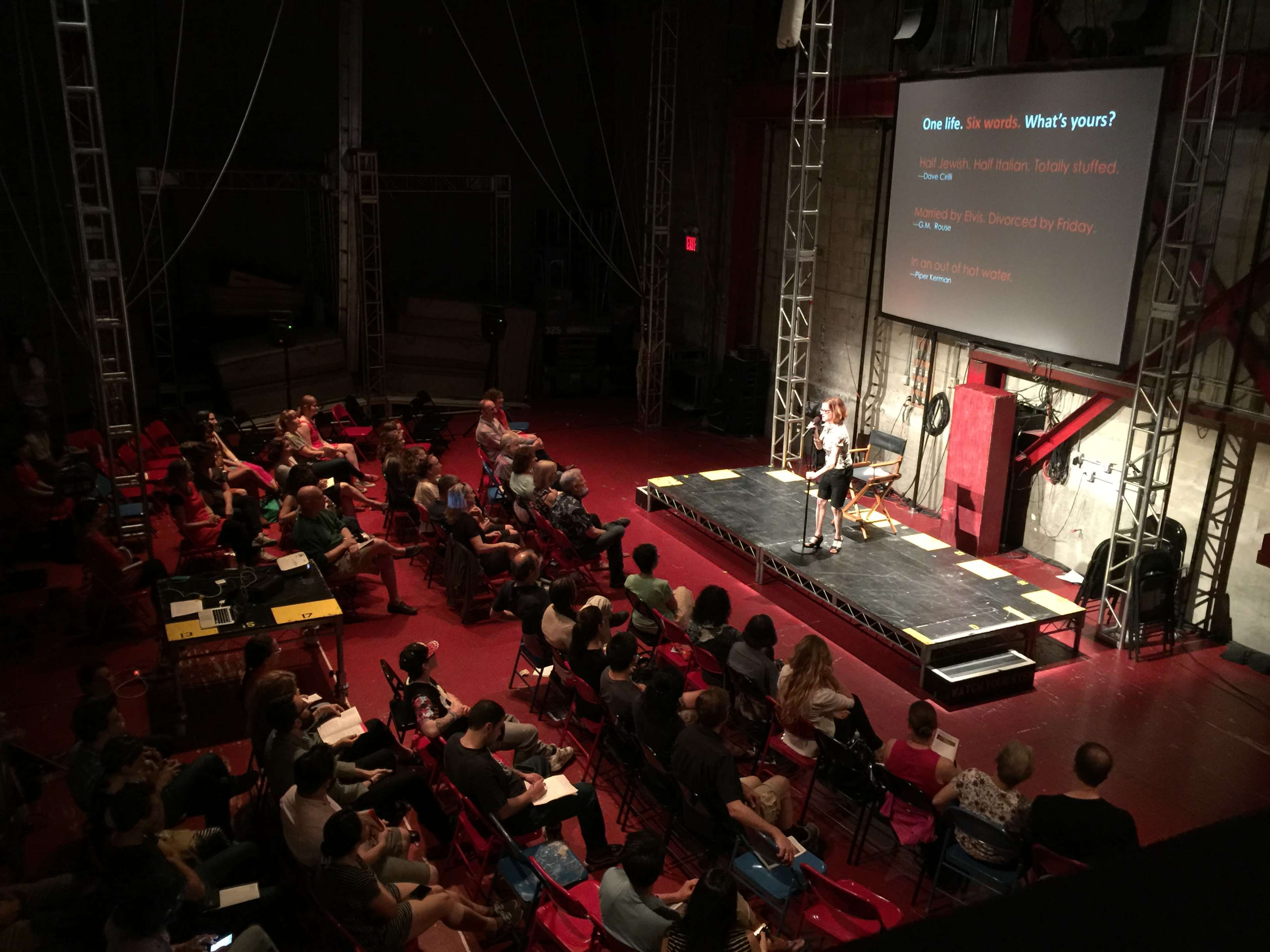 A speaker presents on stage to an audience seated in red chairs in a well-lit auditorium.