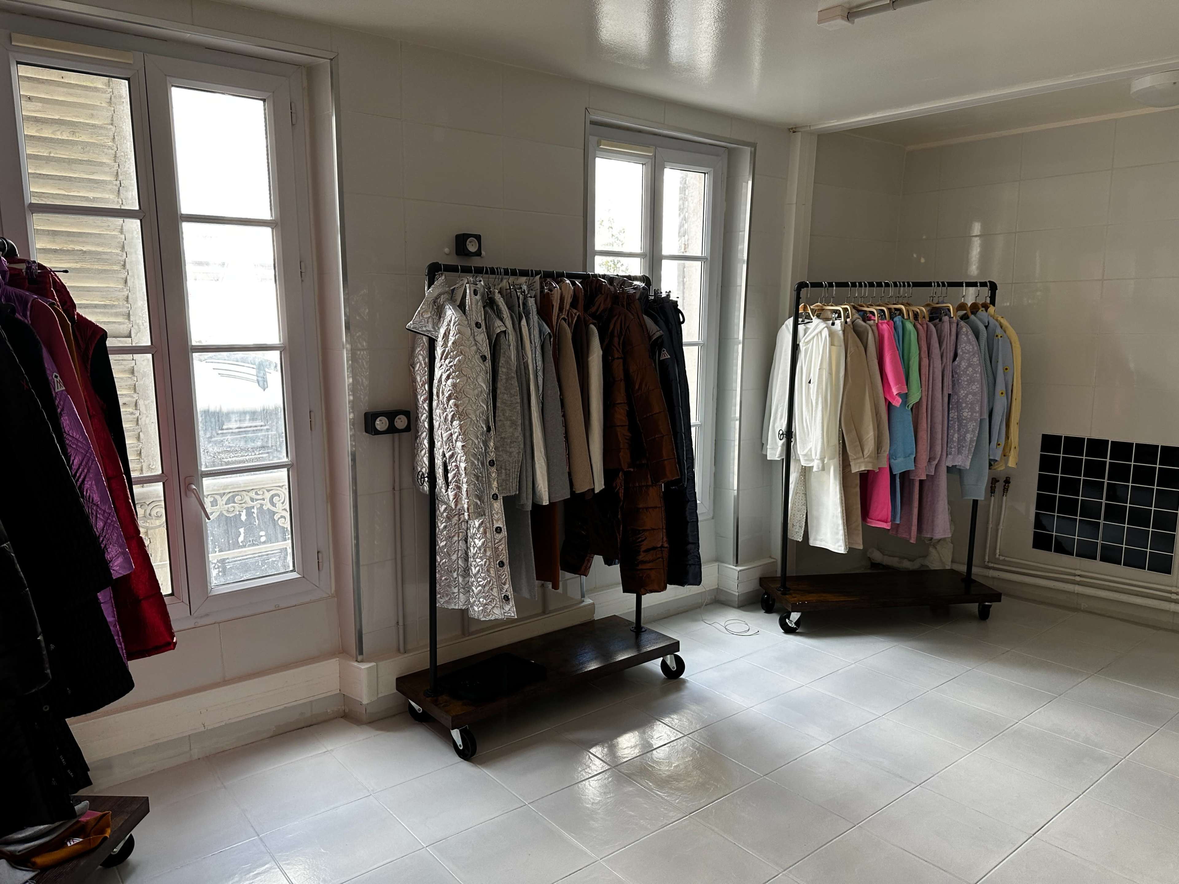 The image shows a room with two clothing racks, one filled with various coats and the other with shirts and dresses, against white tiled walls and large windows.