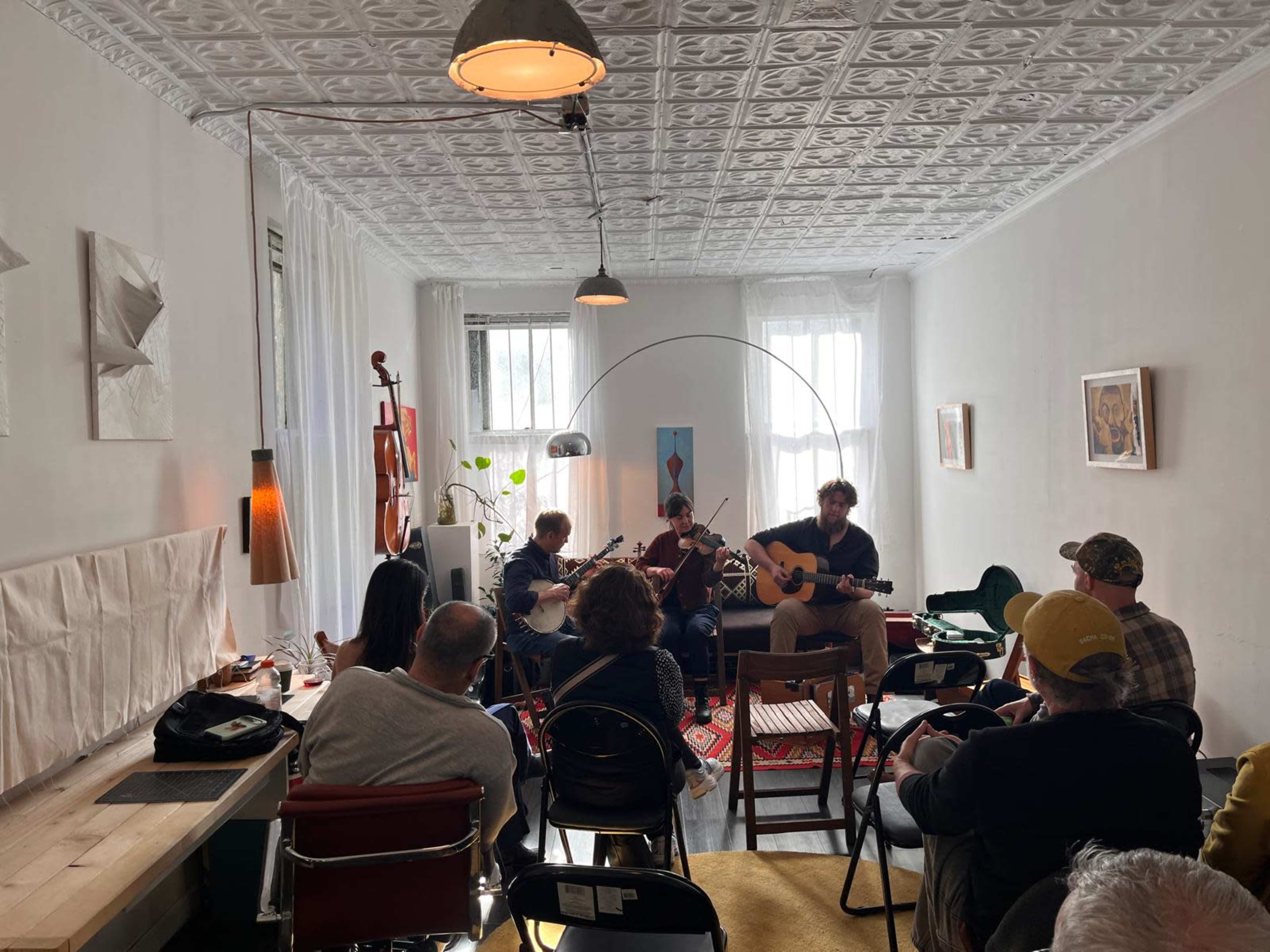 A small crowd sits on folding chairs in a bright room while three musicians perform with guitars and a violin.