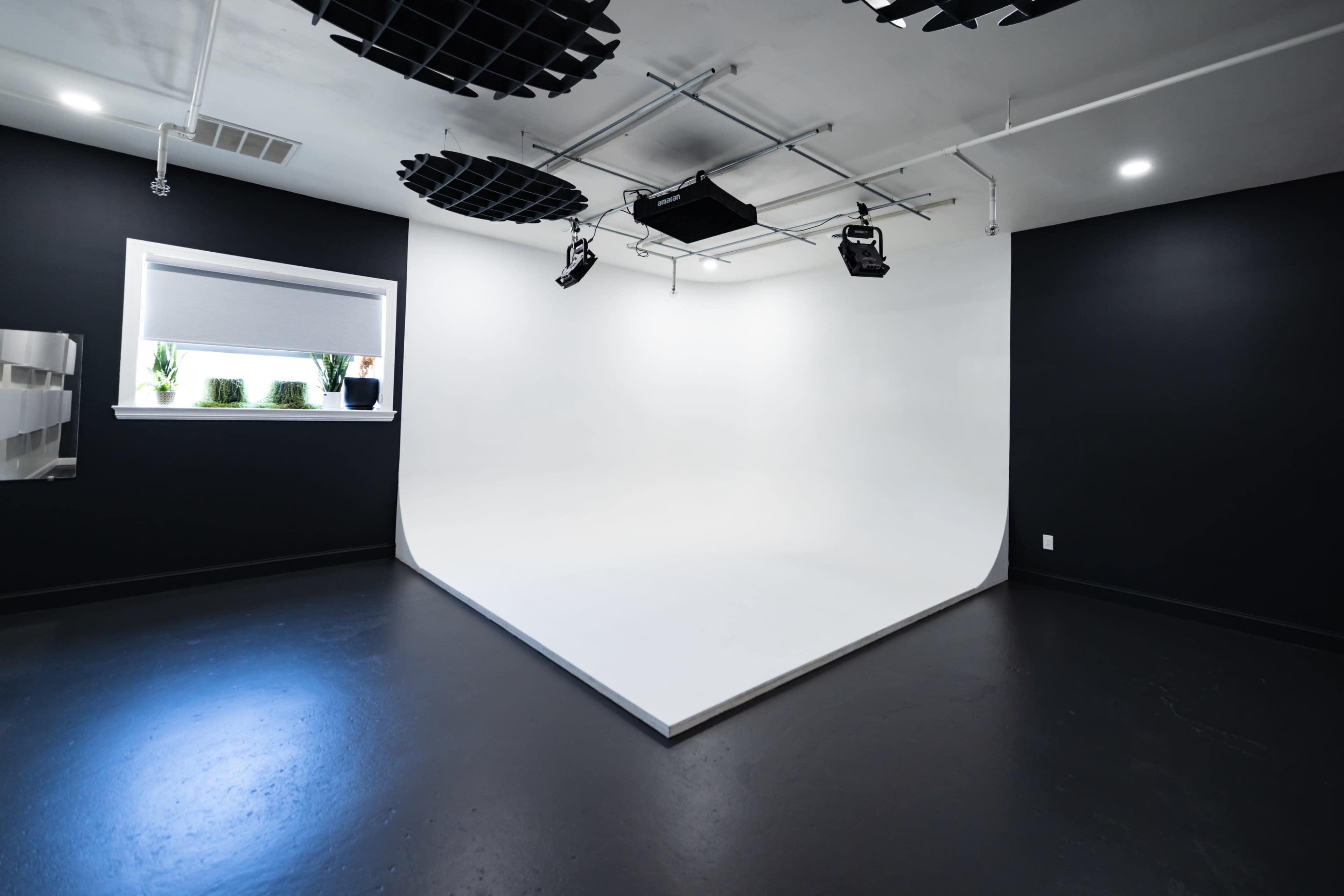 A minimalist studio space with a white cyclorama wall and dark walls, equipped with overhead lighting and a window.