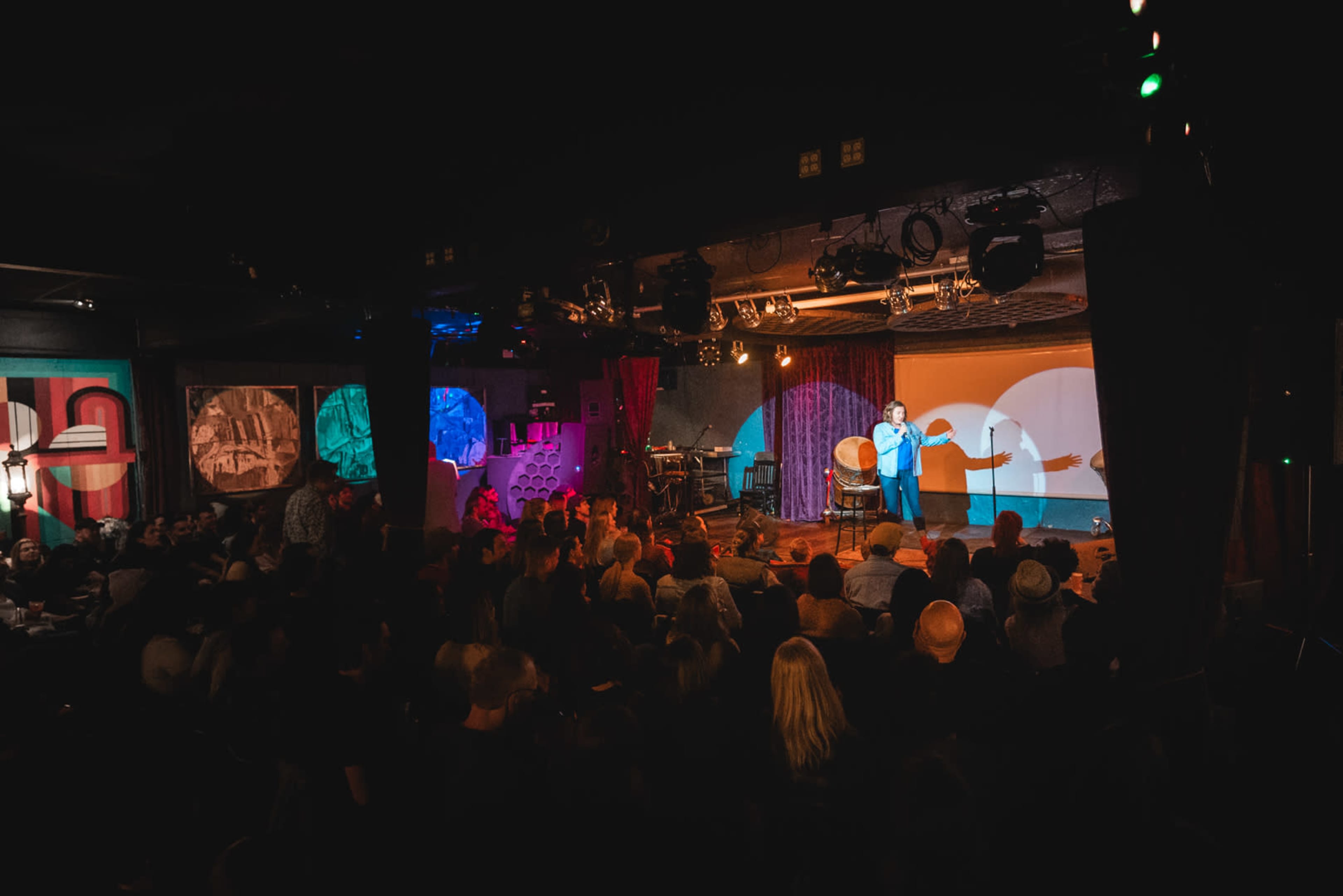 A performer stands on stage in front of a large audience, with colorful projections creating a dynamic backdrop.